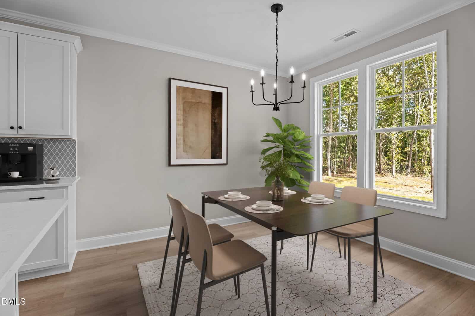A modern dining area with a dark rectangular table, four tan chairs, a plant centerpiece, and a black chandelier. Large windows let in natural light and show trees outside. Neutral walls and minimal art complete the space.