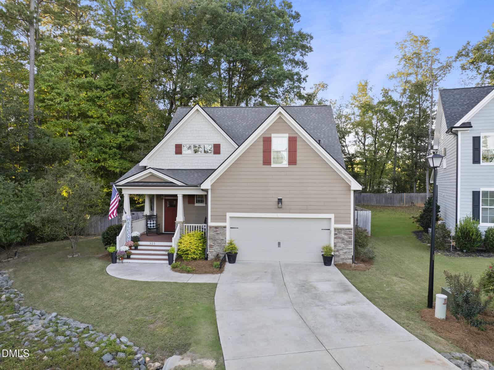 Charming two-story beige house located at 325 Paddy Lane in Youngsville, featuring red shutters, a welcoming front porch, U.S. flag display, and an attached two-car garage with a curved concrete driveway. The well-kept yard boasts lush shrubs and grass, all nestled among trees and neighboring homes.