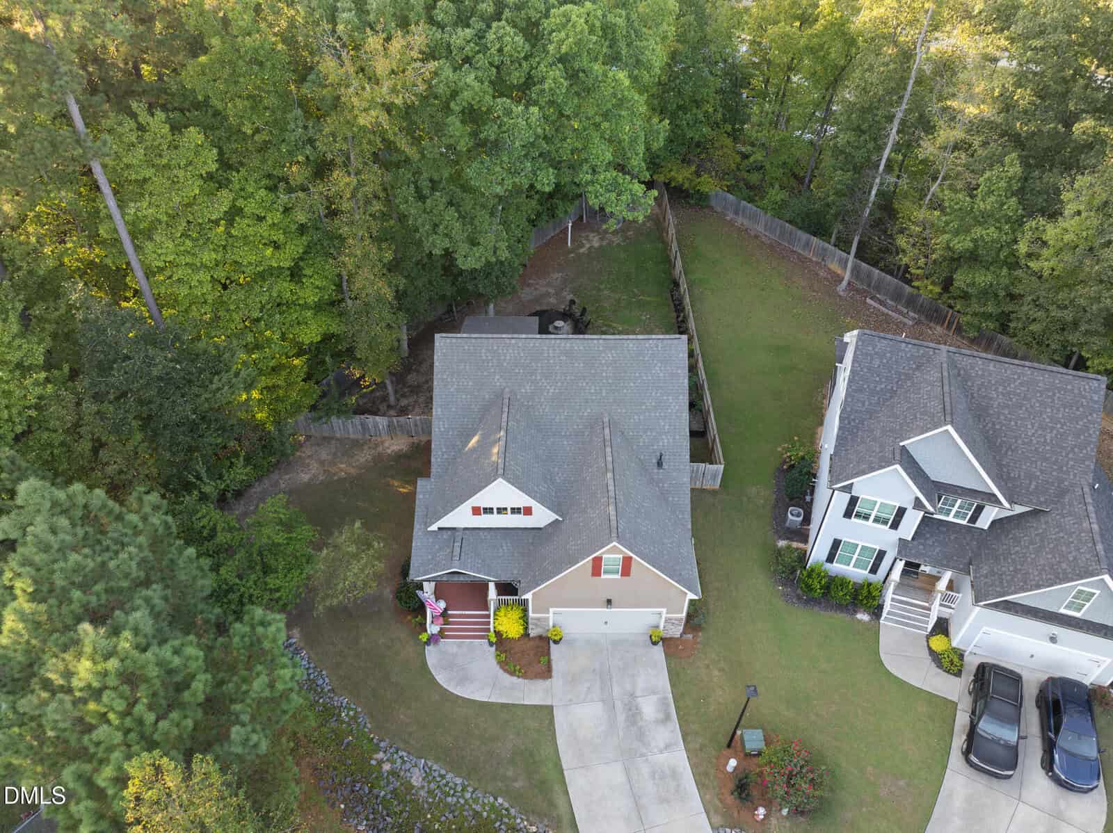 Aerial view of a beige house with a gray roof, a red porch, and a curved driveway. The backyard is fenced and borders a wooded area. A neighboring house and green lawns are visible.