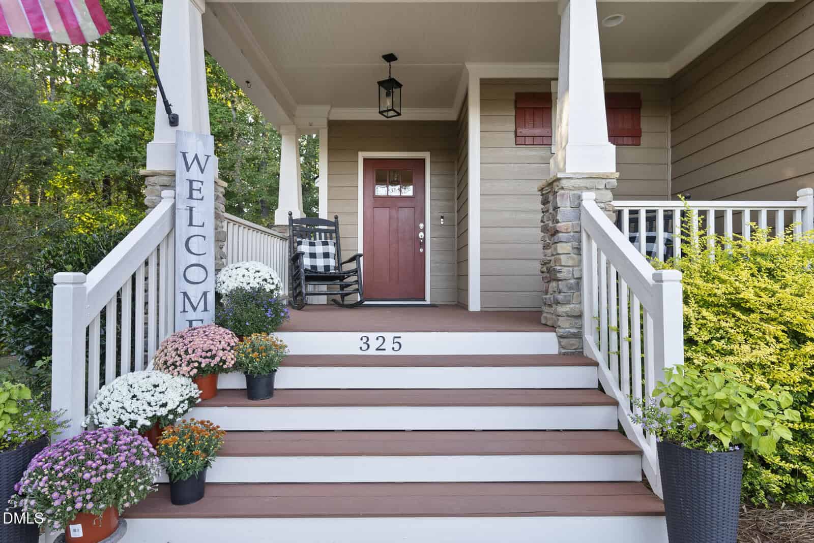 Front porch of a house with a red door, “WELCOME” sign, potted flowers on stairs, and white railings. The house number 325 is painted on the top step. Columns and stone accents frame the entrance.