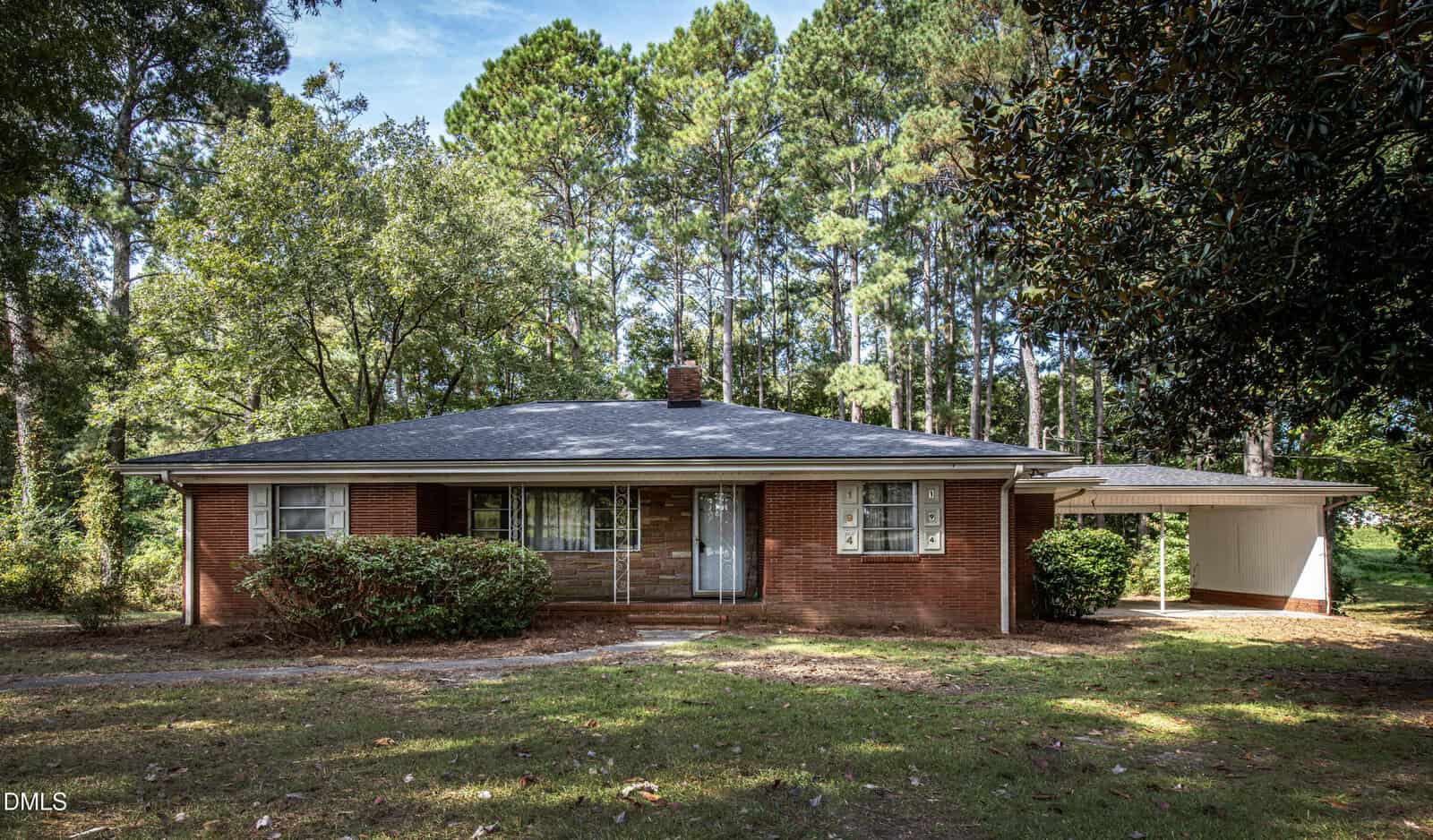Single-story brick house with a dark roof, chimney, front porch, and metal front door, surrounded by bushes and trees on 194 Furney Pearce Road in Zebulon. An open carport is attached to the right side of the house. The lawn is shaded by tall pine trees.