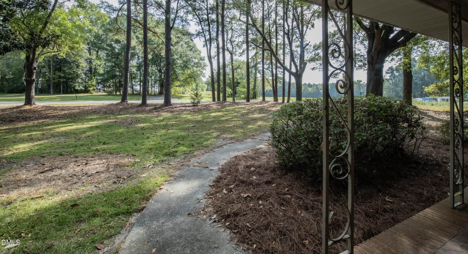A covered porch with decorative metal posts overlooks a curved concrete walkway, shrubbery, and a yard with pine trees and patchy grass. A quiet road runs parallel to the property in the background.