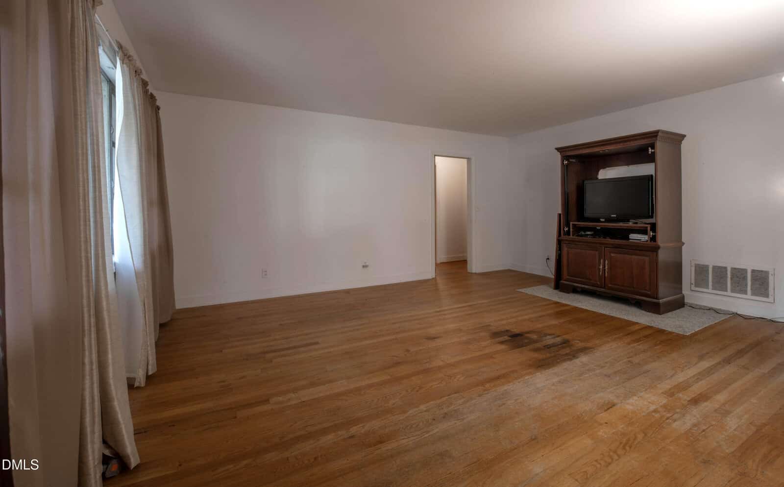 A mostly empty living room with hardwood floors, a large window with curtains, white walls, a doorway, and a wooden TV stand with a television. There is a visible dark stain on the floor near the center.
