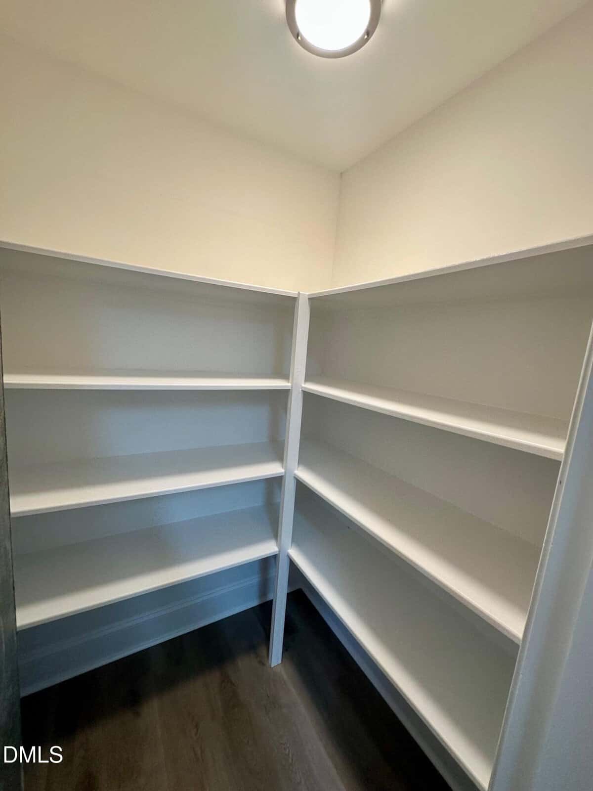 A small walk-in pantry with white built-in shelves on three walls and dark wood flooring, lit by a ceiling-mounted light. The shelves are empty.