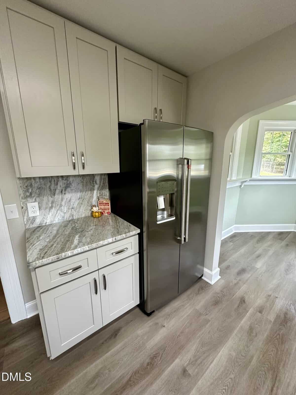 A modern kitchen corner with light-colored cabinets, a stainless steel refrigerator, marble-patterned countertop, and wood-look flooring. An archway opens into a bright room with windows.
