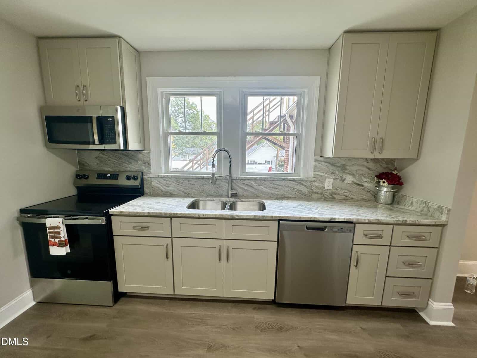 A modern kitchen with light-colored cabinets, marble-patterned countertops, stainless steel appliances including a stove, microwave, and dishwasher, and a large window above a double sink.