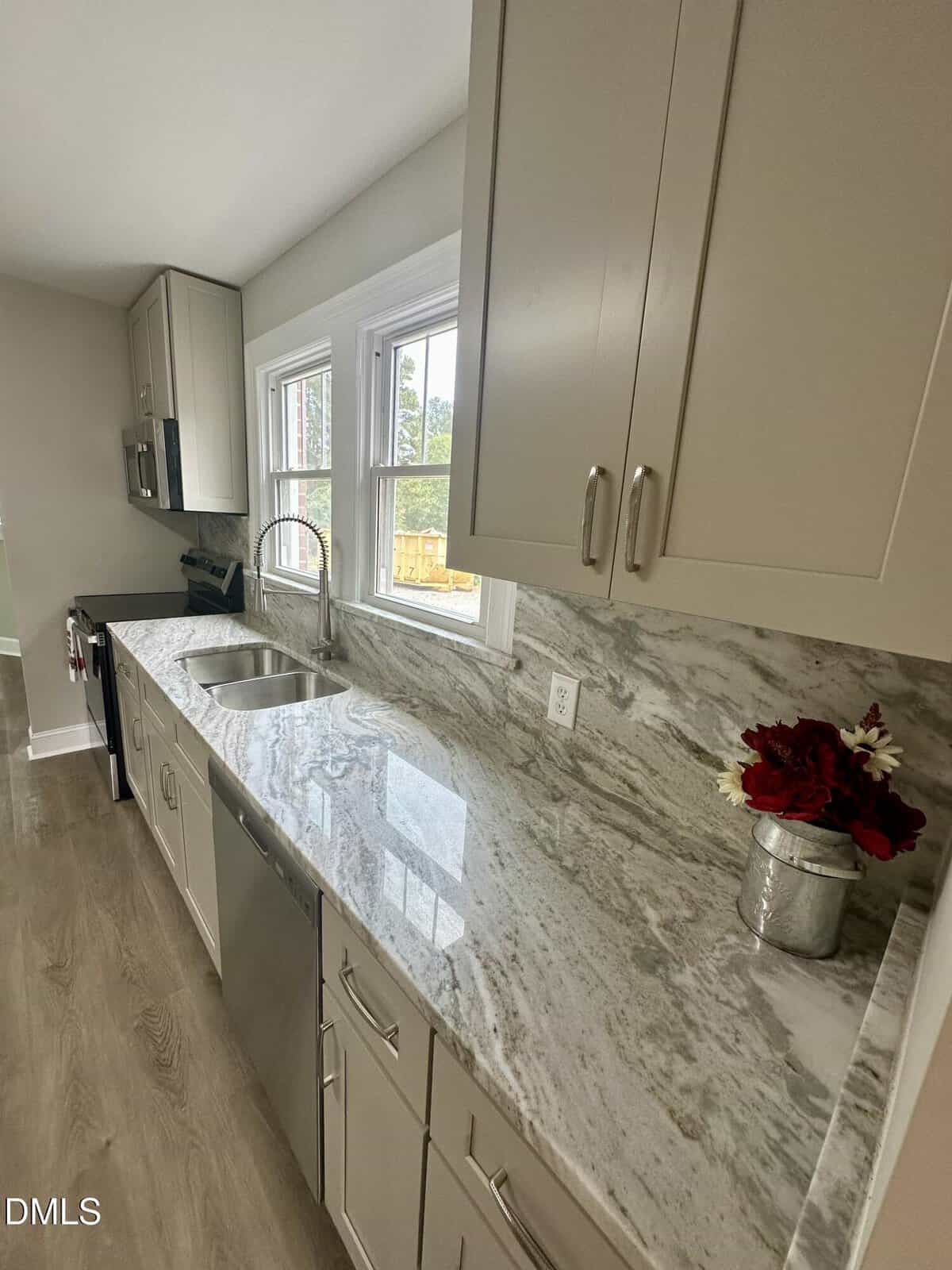 A modern kitchen with light-colored cabinets, a marble-patterned countertop, stainless steel sink and appliances, and a small metal vase with red flowers placed on the counter near the window.