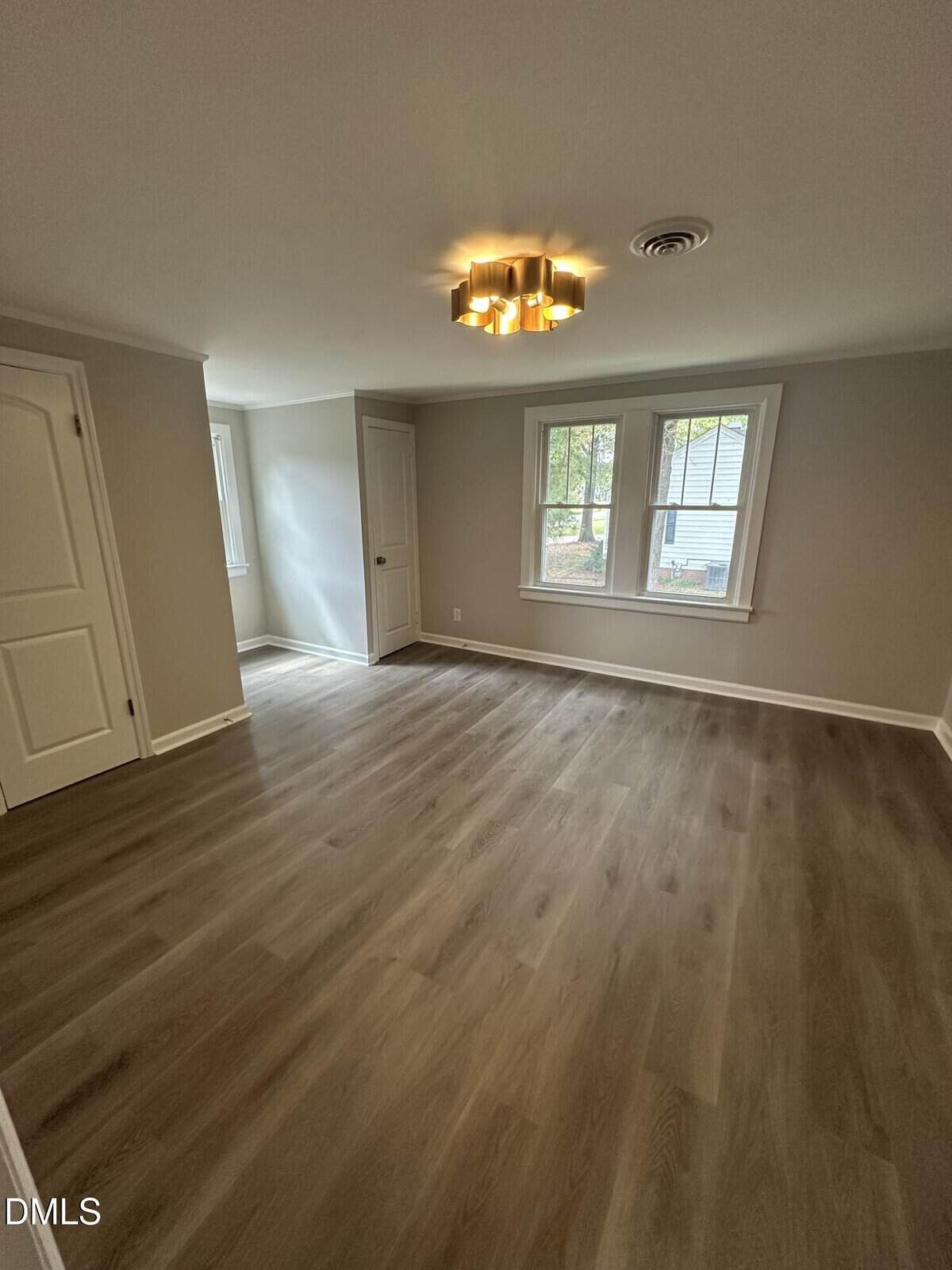 Empty room with light wood flooring, beige walls, two windows, a modern ceiling light fixture, and two white doors. Natural light comes through the windows, creating a bright and spacious atmosphere.