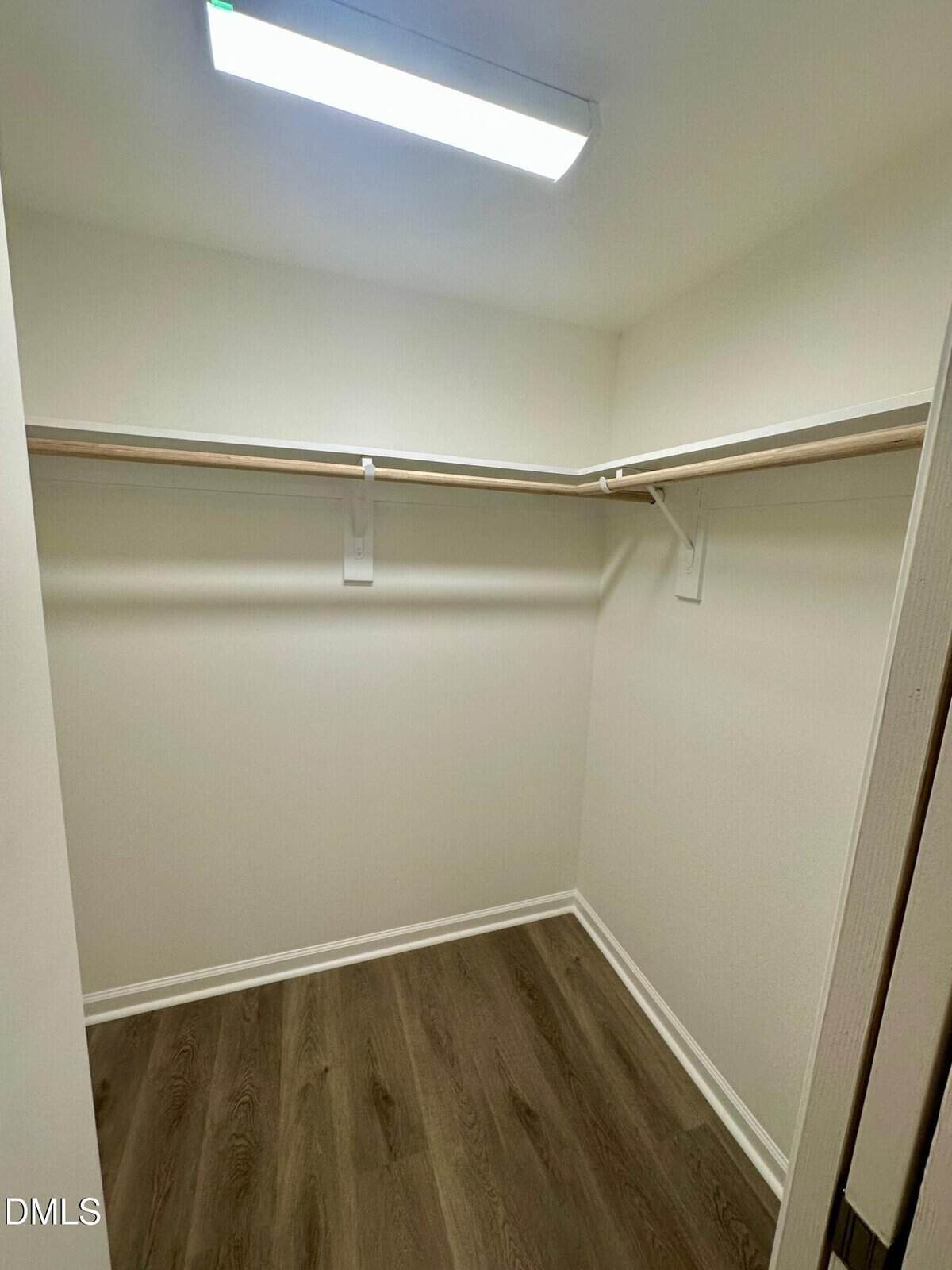Empty walk-in closet with white walls, a wooden floor, two wooden hanging rods with shelves on adjacent walls, and a ceiling-mounted fluorescent light fixture.