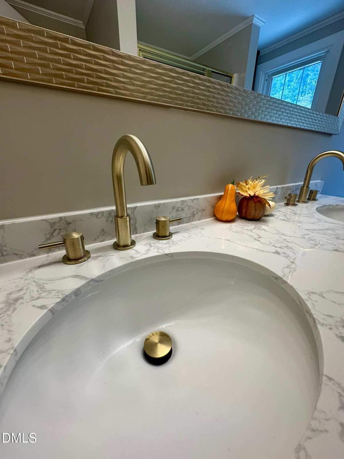 A bathroom countertop with a marble pattern features a gold faucet and handles. A small arrangement of decorative gourds and artificial flowers sits near the wall beneath a large mirror.