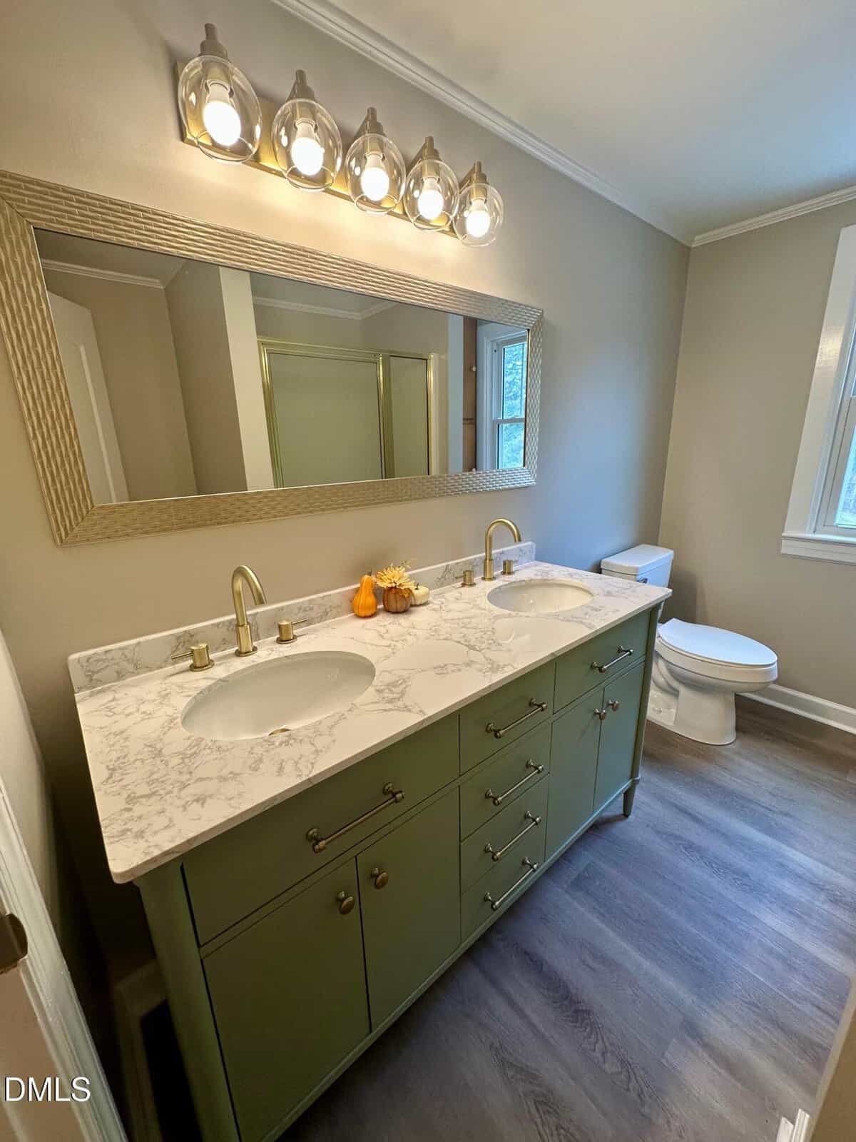A bathroom with a green double-sink vanity, marble countertop, brass fixtures, a large mirror, overhead lighting with five bulbs, a white toilet, wood-look flooring, and beige walls.