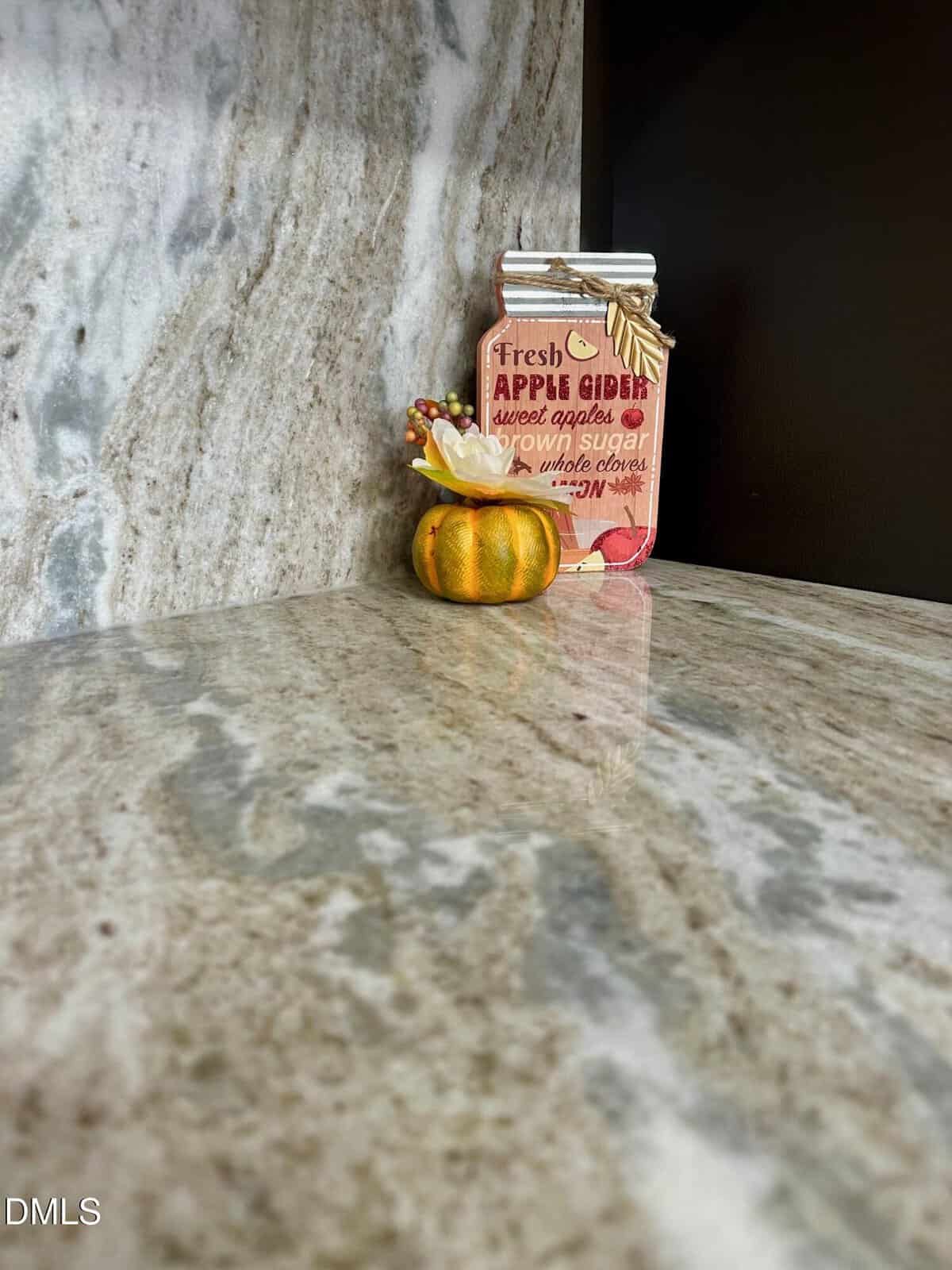 A small decorative pumpkin and a jar-shaped sign reading "Fresh Apple Cider" sit on a polished marble countertop against a marble-patterned backsplash.