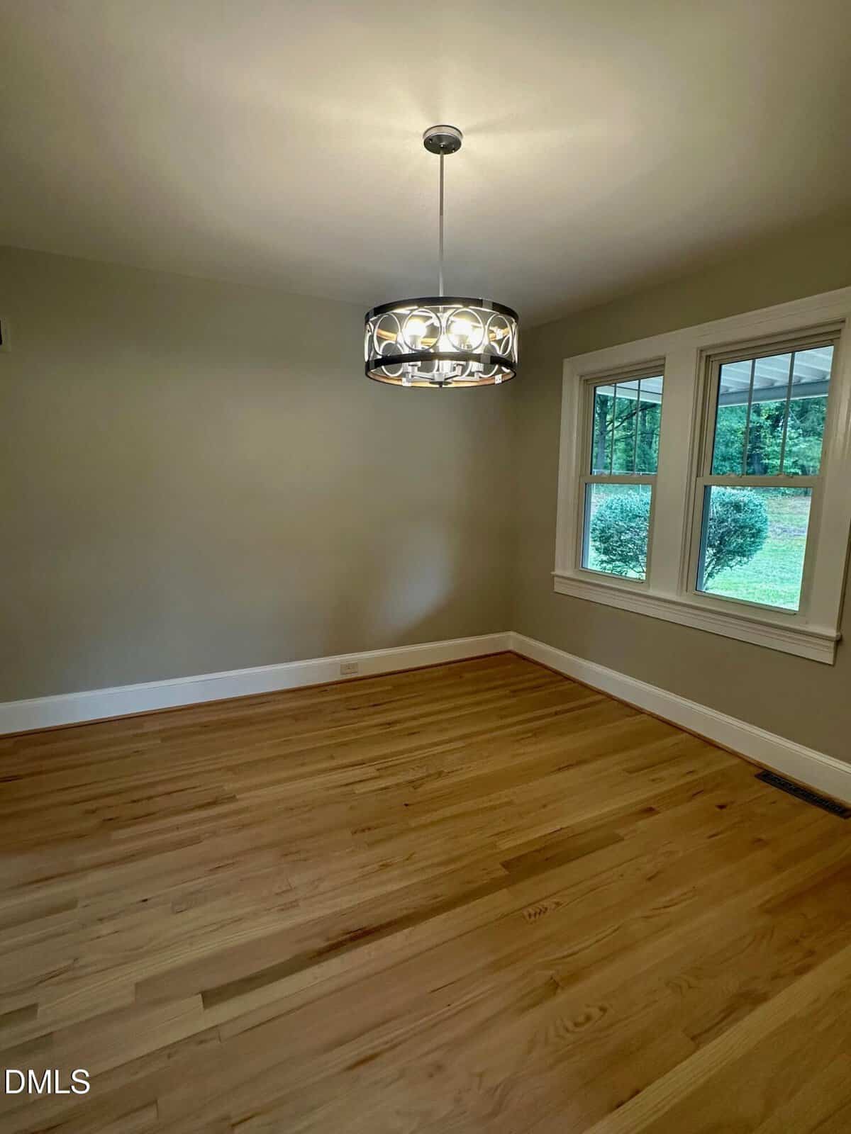 Empty room with light wood floors, beige walls, a modern circular ceiling light fixture, and two large windows showing greenery outside.