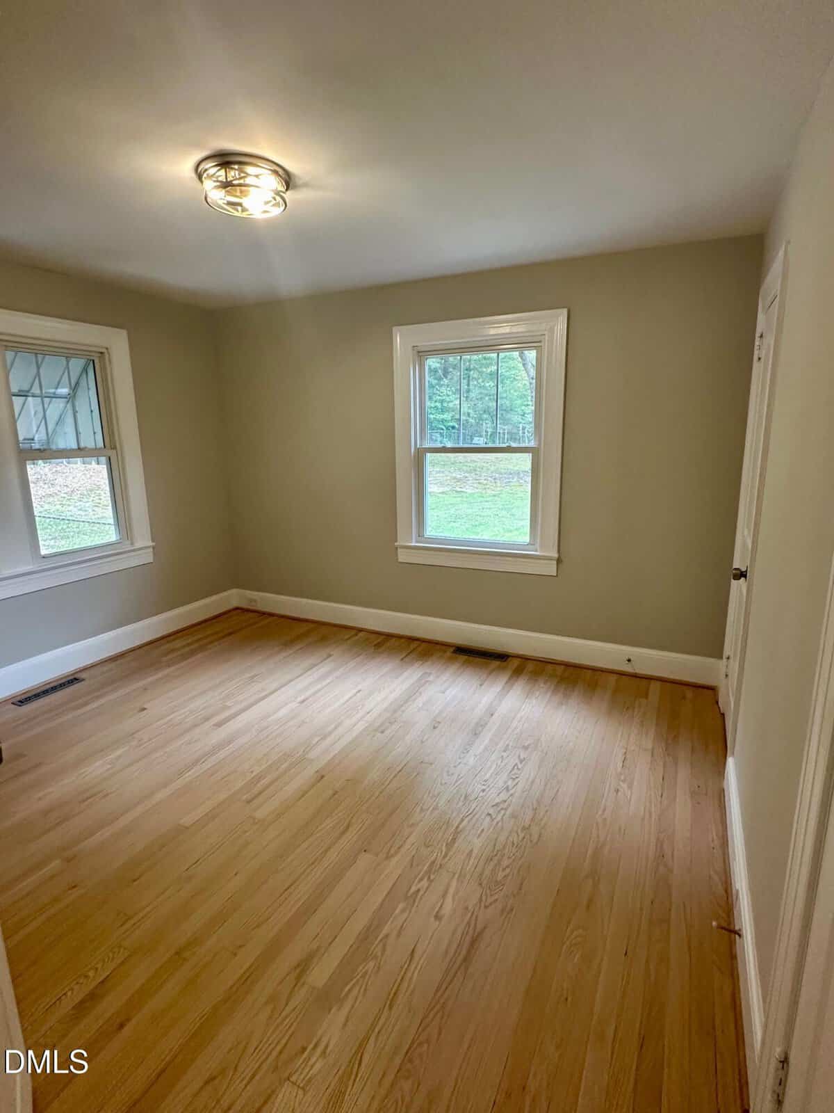 A small empty room with light wood floors, beige walls, two windows with white trim, a white ceiling, and a ceiling light fixture. Natural light brightens the space.