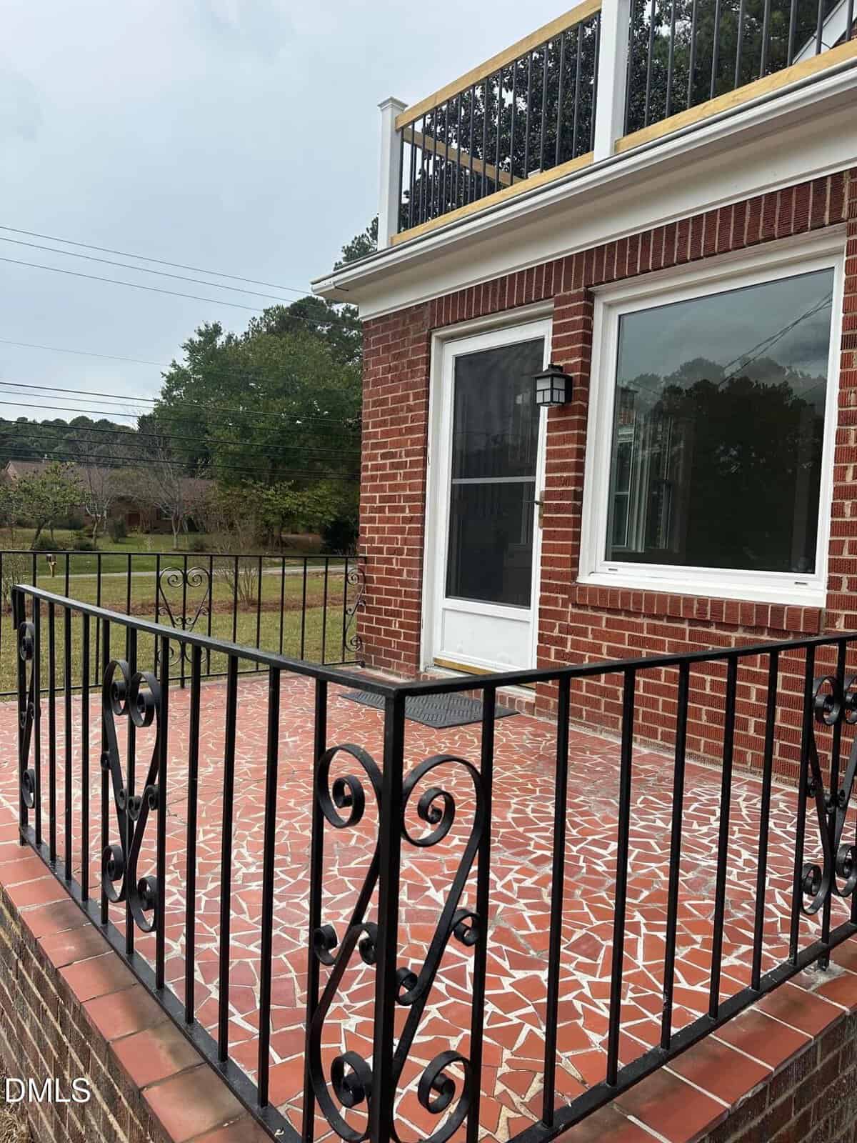A red brick house with a white-framed glass door leads to a small patio with a red and white tile floor. The patio is enclosed by a black metal railing and overlooks a grassy yard with trees in the background.
