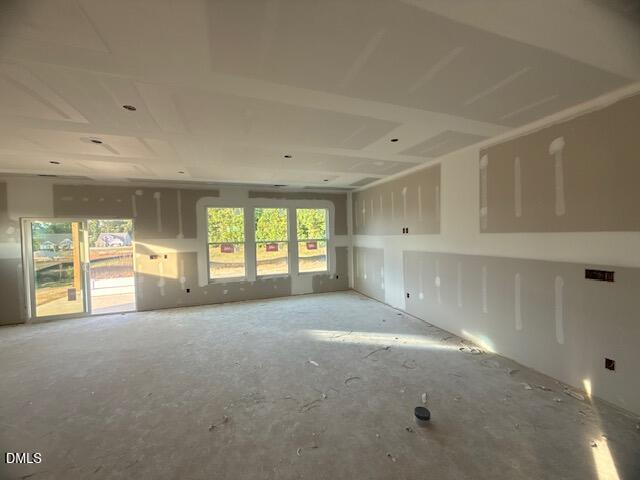 Unfinished room with drywall installed, unpainted walls, exposed electrical outlets, and three large windows. Sunlight shines in from the windows and glass door, casting shadows on the bare concrete floor.