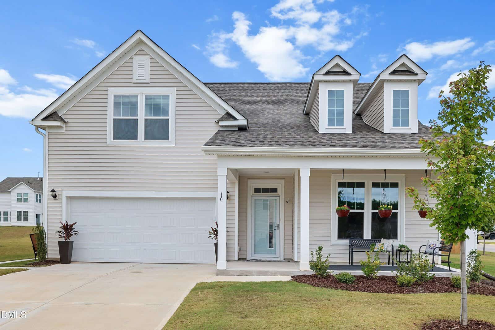 A two-story house with light gray siding, a covered front porch, double garage, and dormer windows. The yard has grass, mulch, a small tree, plants, and a driveway under a partly cloudy blue sky.
