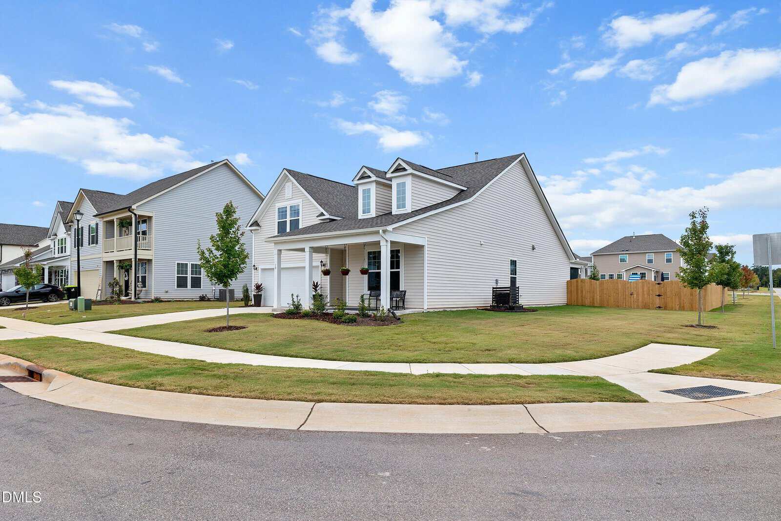 A white, single-story house with a covered front porch and attached garage sits on a corner lot with a neatly maintained lawn and young trees. Sidewalks curve around the property under a blue sky with scattered clouds.