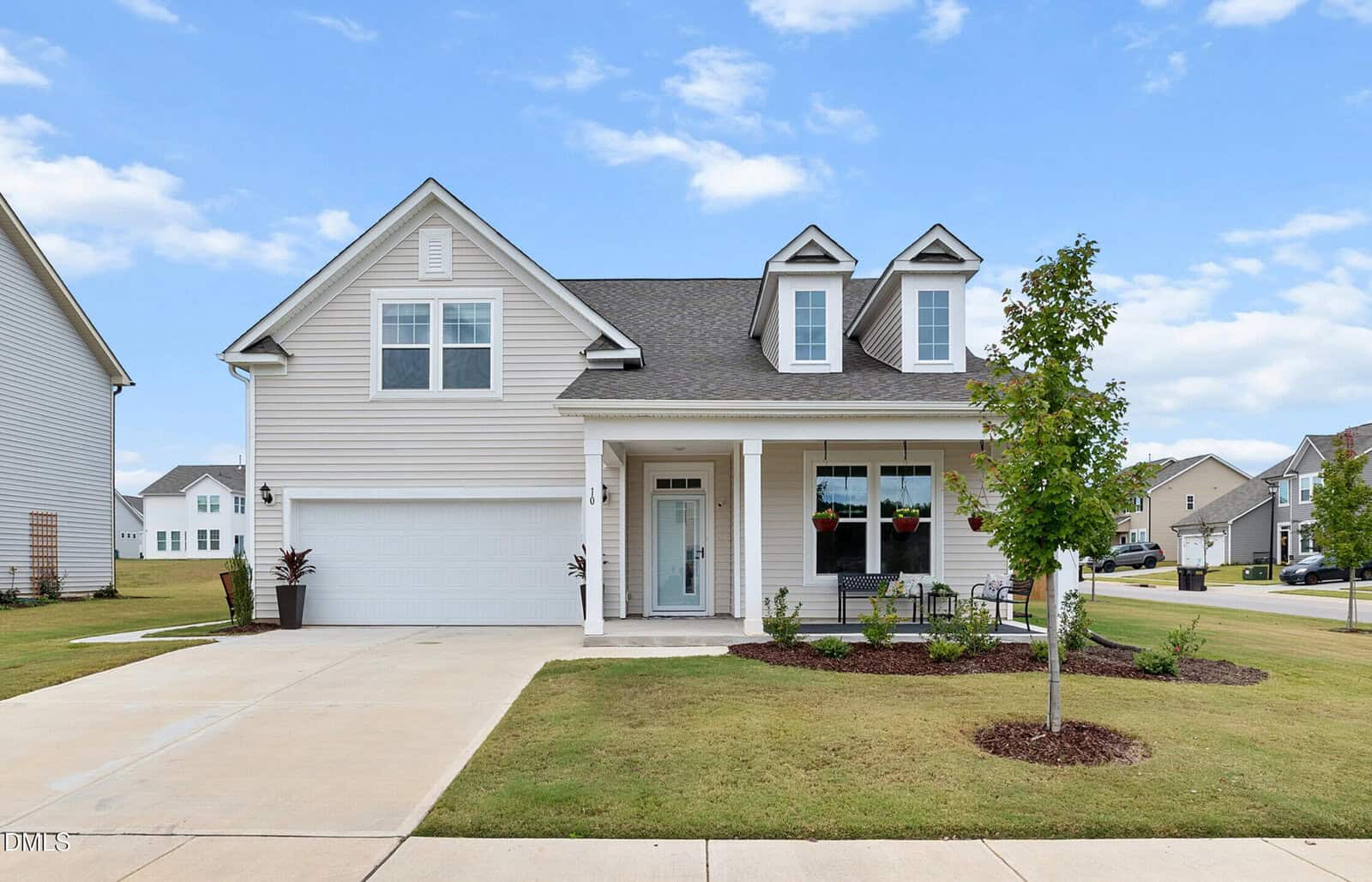 A modern two-story house with beige siding, a double garage, front porch, and dormer windows sits on a corner lot at 10 Grant Mercer Drive in Franklinton. The yard features trimmed grass, a young tree, and minimal landscaping. Nestled in a suburban neighborhood, this home blends comfort and curb appeal.