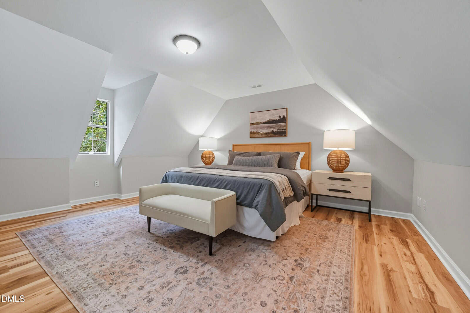 A bedroom with sloped ceilings features a bed with gray and white bedding, two nightstands with lamps, a cushioned bench at the foot of the bed, a patterned rug, wood flooring, and a single window.