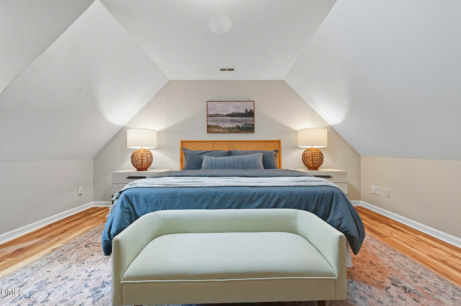 A cozy attic bedroom with sloped ceilings, a bed with blue bedding, two nightstands with lamps, a painting above the bed, a bench at the foot, and wood floors.
