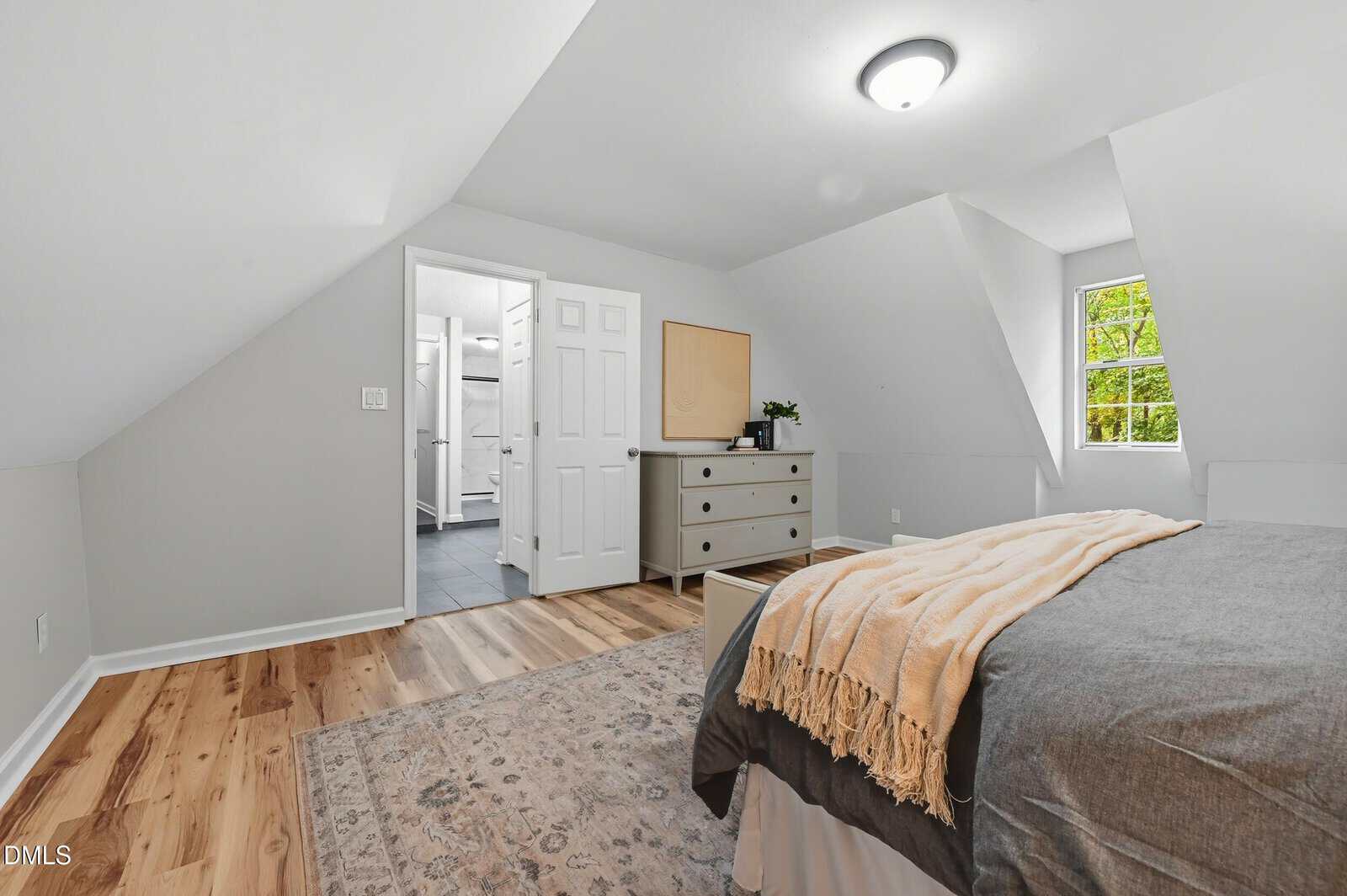 Bright attic bedroom with sloped ceiling, bed with beige throw, dresser, rug on wooden floor, small window, and open door to a bathroom.
