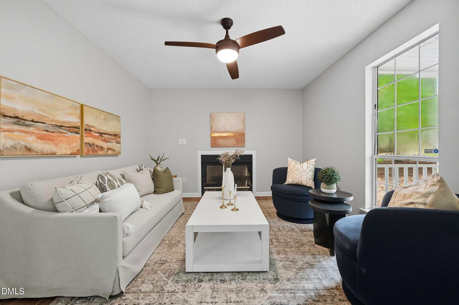 A living room with a white sofa, white coffee table, two dark blue armchairs, and a fireplace. Neutral artwork hangs above the sofa and fireplace. There is a ceiling fan and a large window on the right.