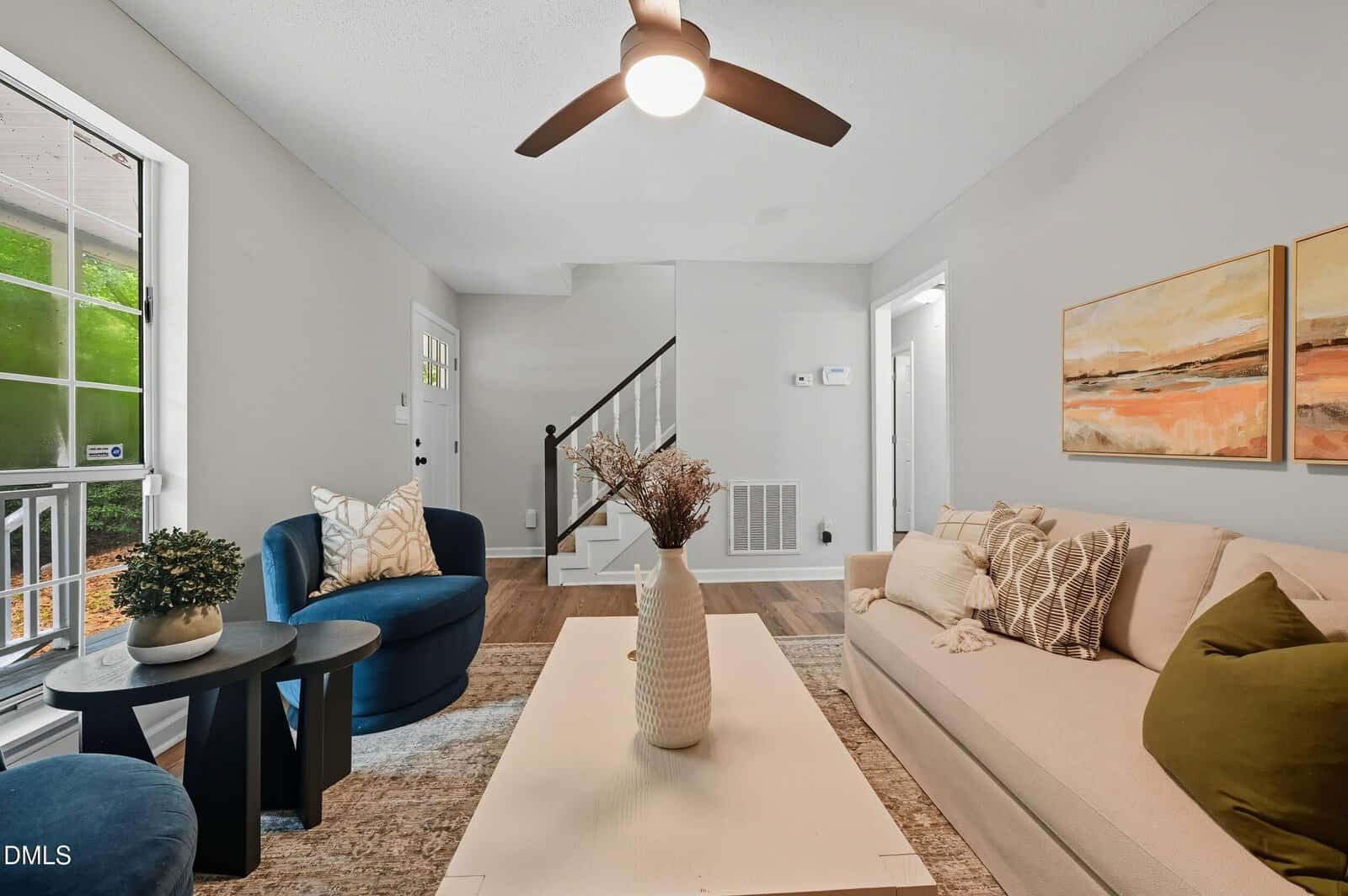 A modern living room with neutral walls, a beige sofa with cushions, a blue armchair, a white coffee table with a vase, abstract wall art, a ceiling fan, and large window letting in natural light.