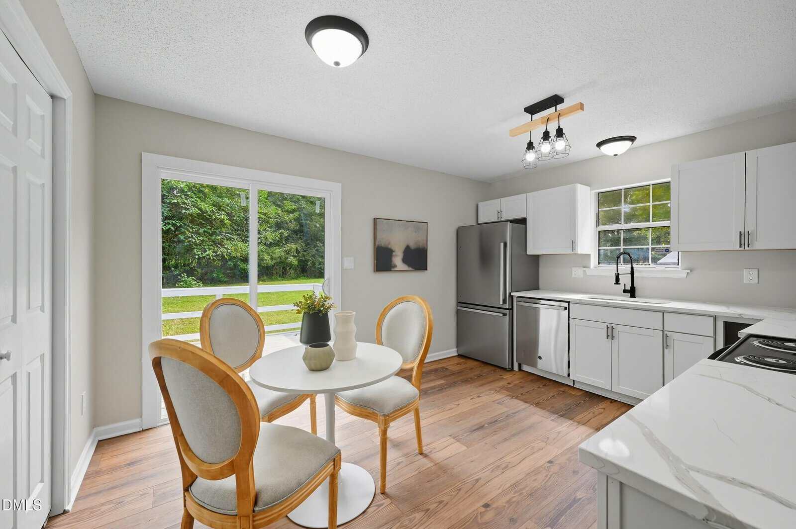 A modern kitchen with white cabinets and marble countertops, featuring a round dining table with four chairs, stainless steel appliances, wood flooring, and a large sliding glass door opening to a grassy yard.