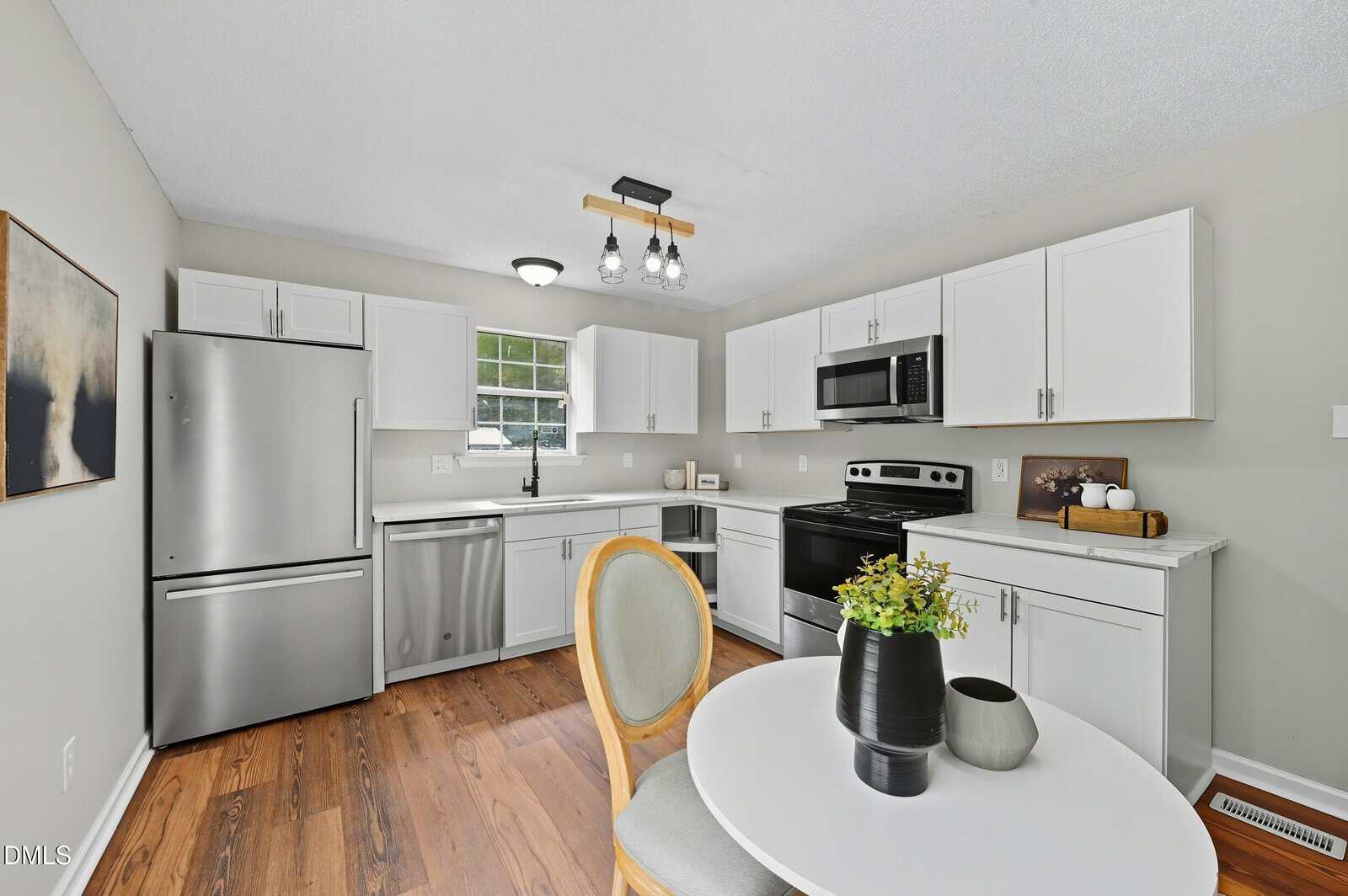 A modern kitchen with white cabinets, stainless steel appliances, and hardwood floors. A round table with chairs and a plant centerpiece is in the foreground. A small window provides natural light above the sink.