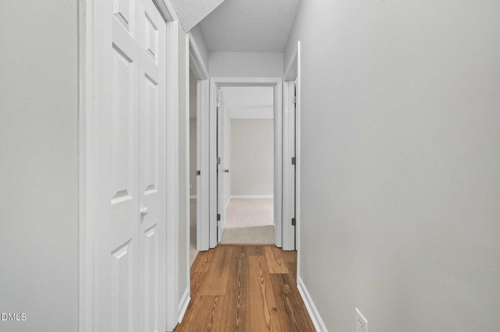 A hallway with light gray walls, white trim, and wooden flooring. There are closed white doors on the left and open doorways at the end and right side, leading to carpeted rooms.