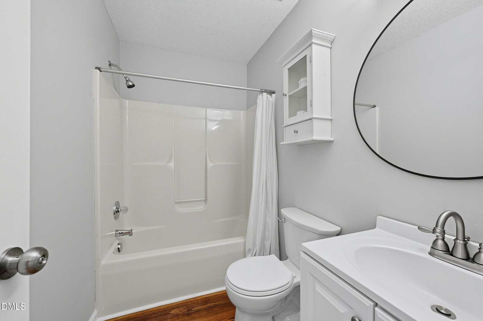 A small bathroom with a white bathtub and shower, a white toilet, a white vanity with a sink and silver faucet, a wall cabinet, a round mirror, and light gray walls. The floor appears to be wood or wood-look.