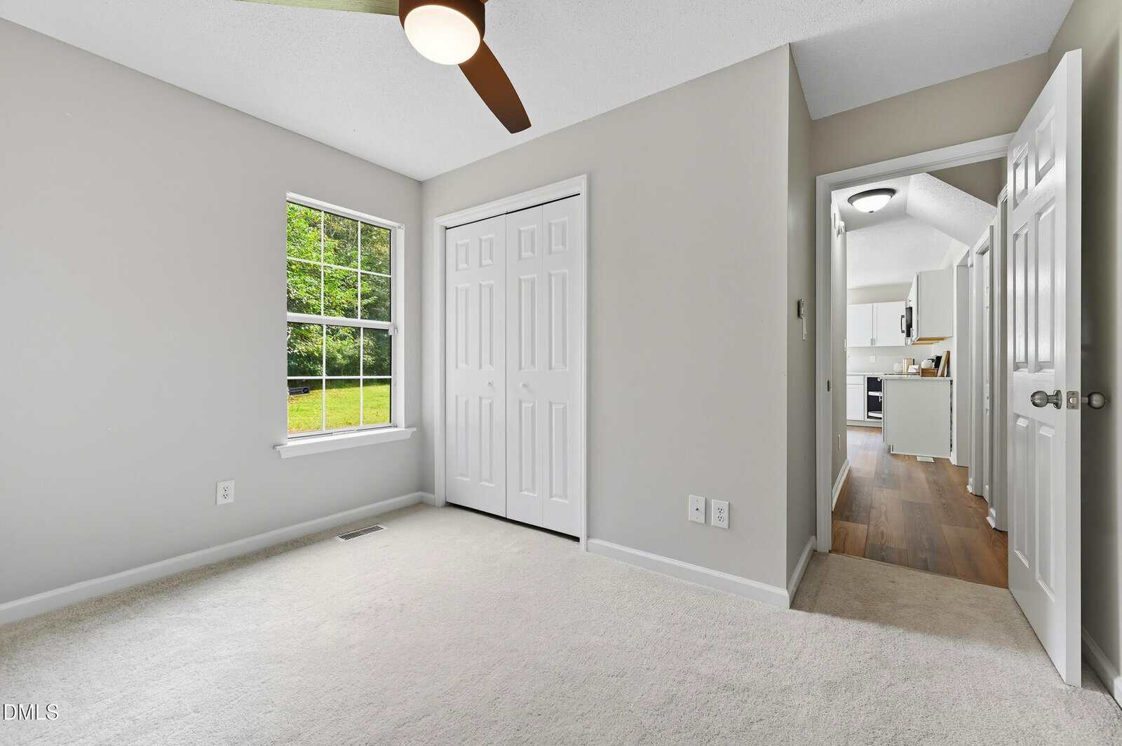 A small bedroom with light-colored walls and carpet, a window showing green grass outside, a ceiling fan, a closed closet with double doors, and an open door leading to a hallway and kitchen area.