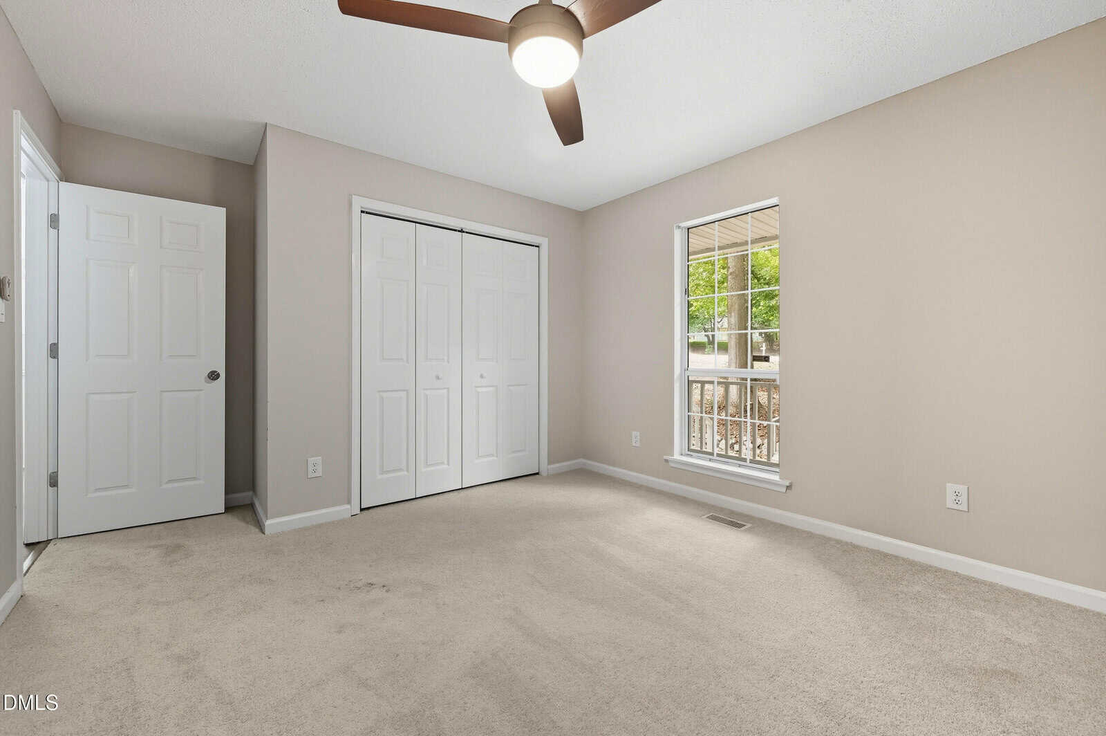 A carpeted, empty bedroom with beige walls, a ceiling fan with a light, a window with a view of a porch, a closet with double doors, and an open door leading to another room.