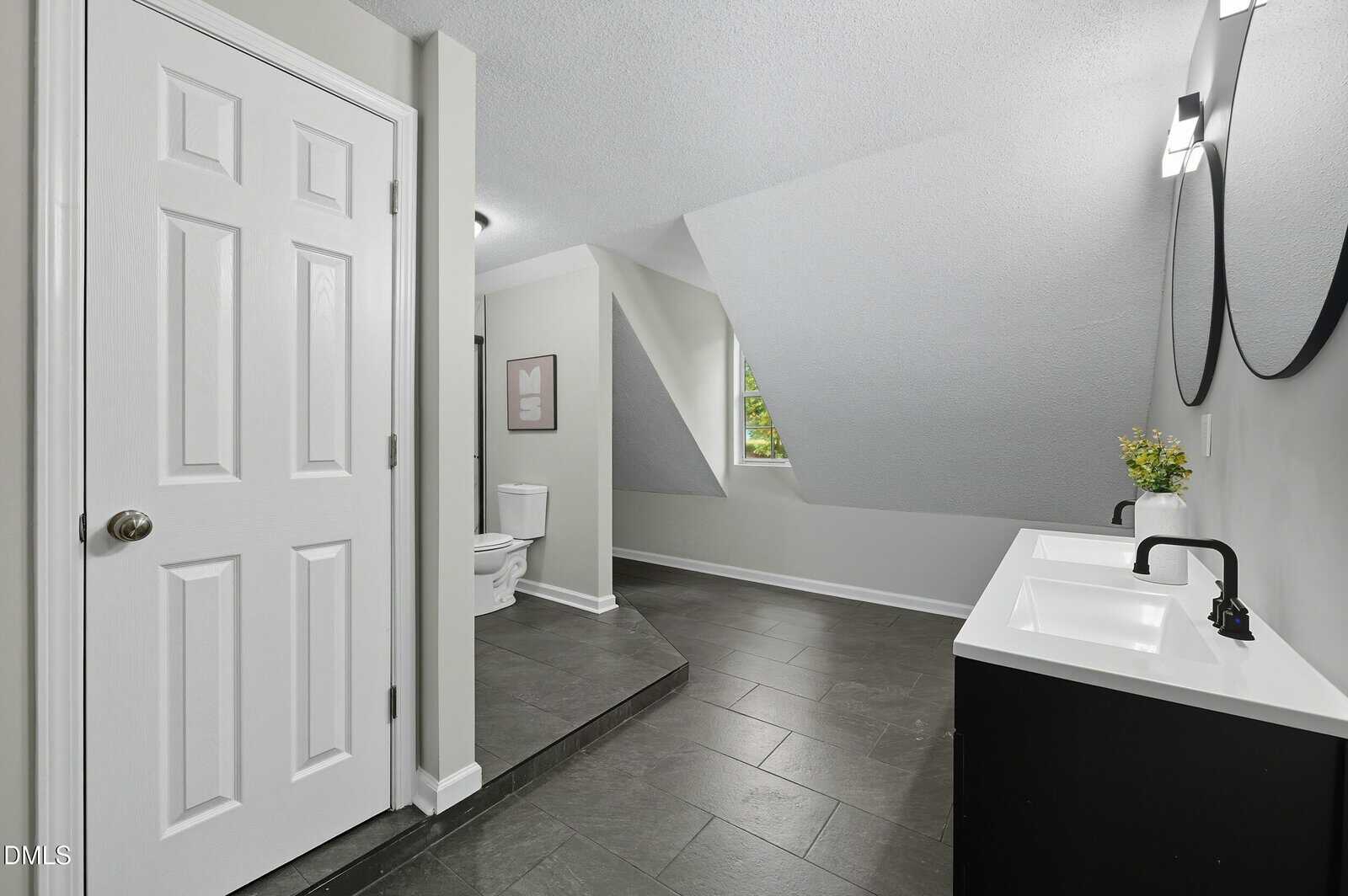 Modern bathroom with black floor tiles, a double sink vanity with two mirrors, a toilet in the corner, sloped ceiling, a window with greenery outside, and a white door on the left. Minimalist decor with a small plant.