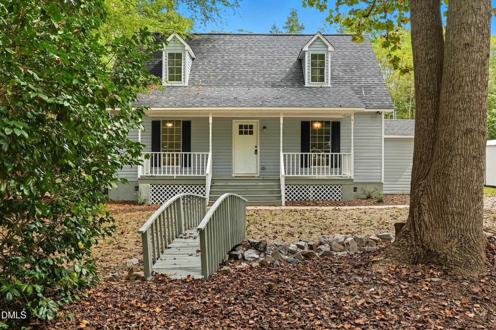 A gray two-story house at 109 Shearin Court, Youngsville, features dormer windows, a covered front porch with black shutters and a white door. A small wooden footbridge crosses rocky terrain, surrounded by trees and shrubs in the yard.
