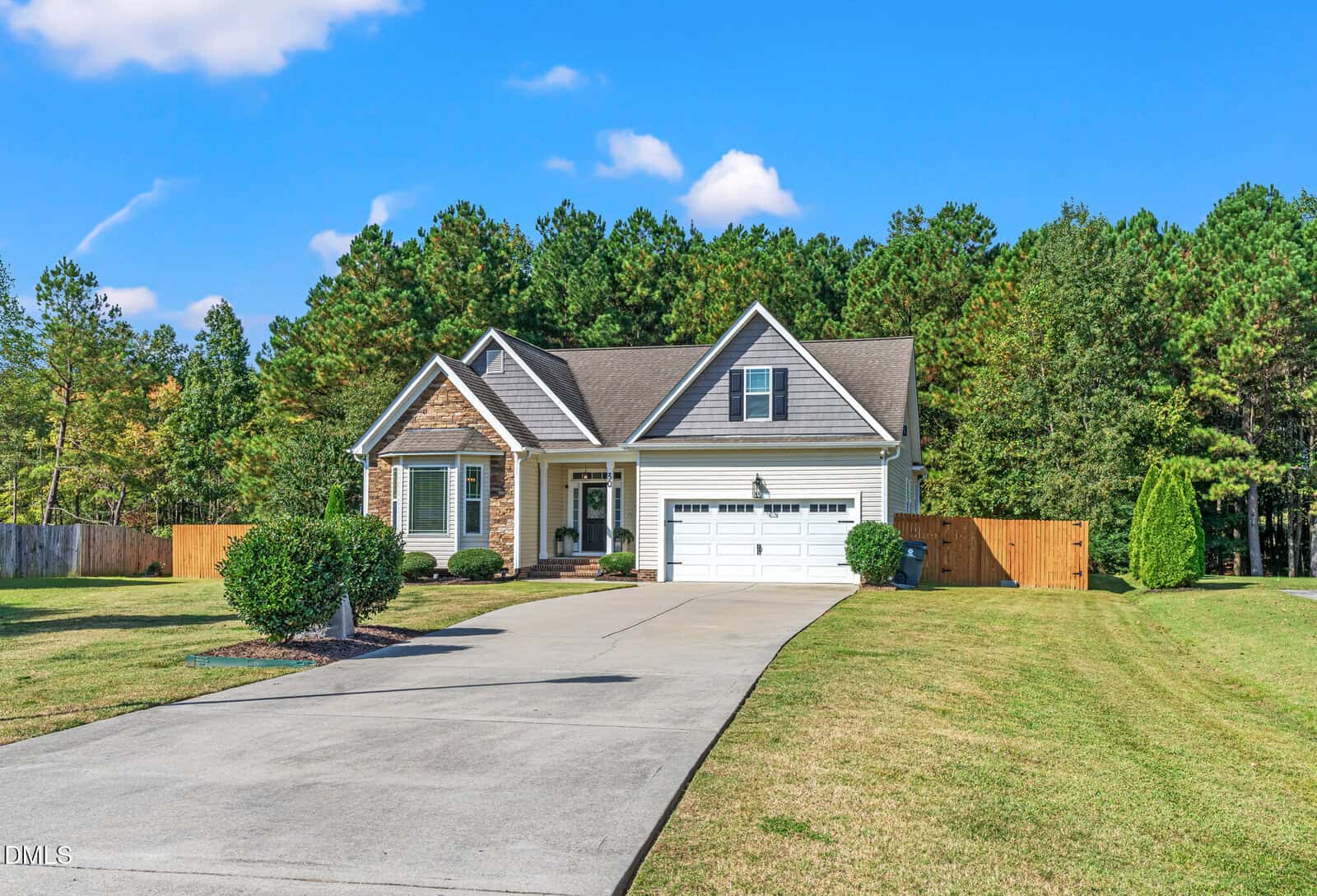 Single-story house with a front porch, double garage, and gabled roof situated in peaceful Youngsville. The driveway at 30 Kinderkamack Rd. Road curves through a well-kept lawn, while the property is bordered by a wooden fence and trees in the background under a blue sky with clouds.