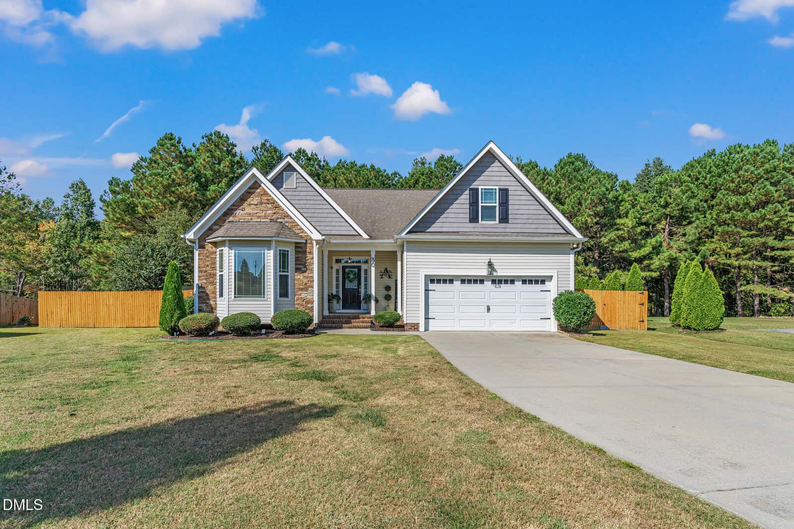Single-story house with a gabled roof, stone and gray siding, two-car garage, manicured lawn, and a long driveway. The home is set against a backdrop of trees under a blue sky with scattered clouds.