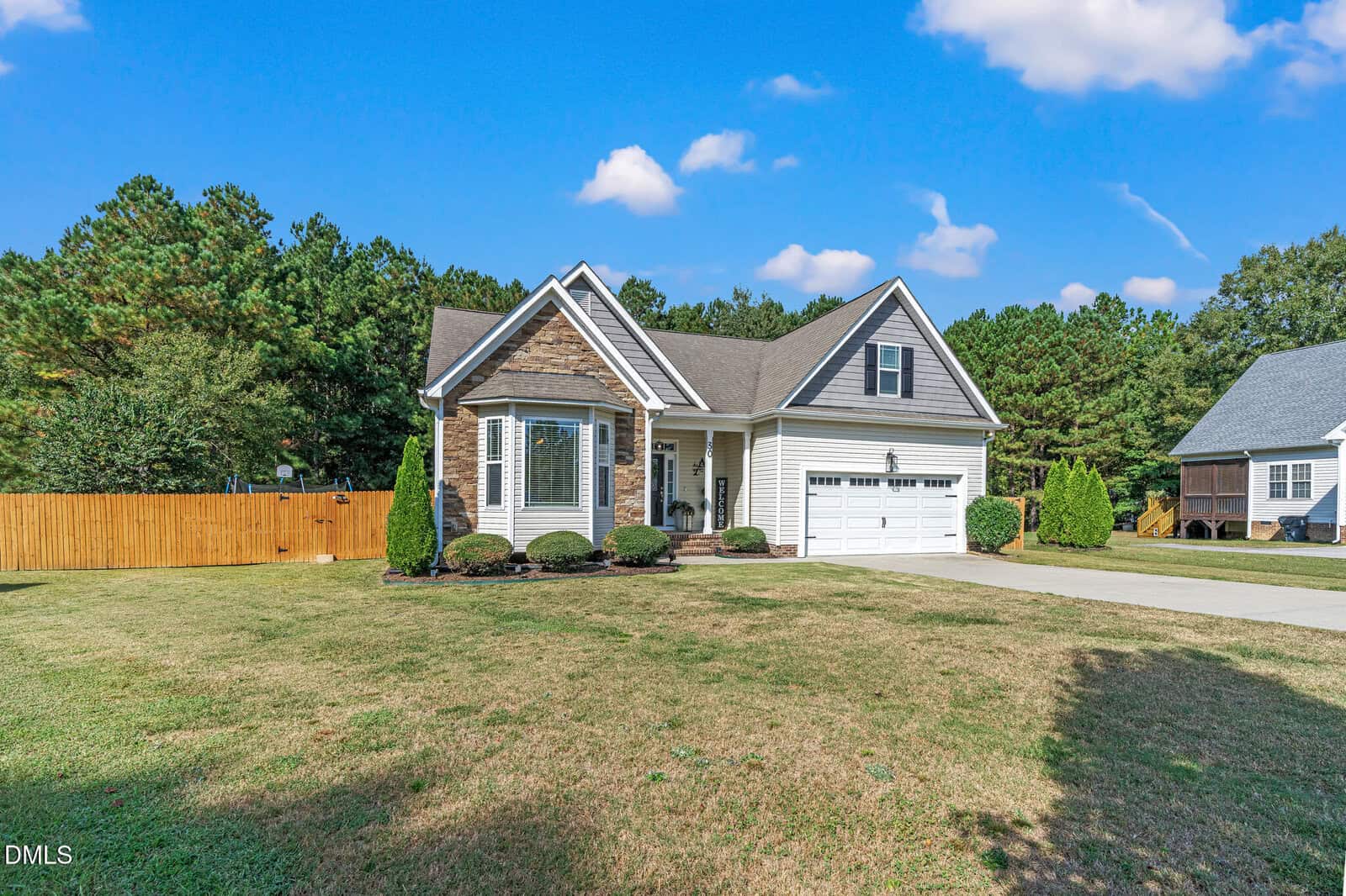 Single-story house with gray siding, stone accents, white trim, and attached two-car garage. Concrete driveway, manicured bushes, large front lawn, and wooden fence on the left. Pine trees and a neighboring home in the background.
