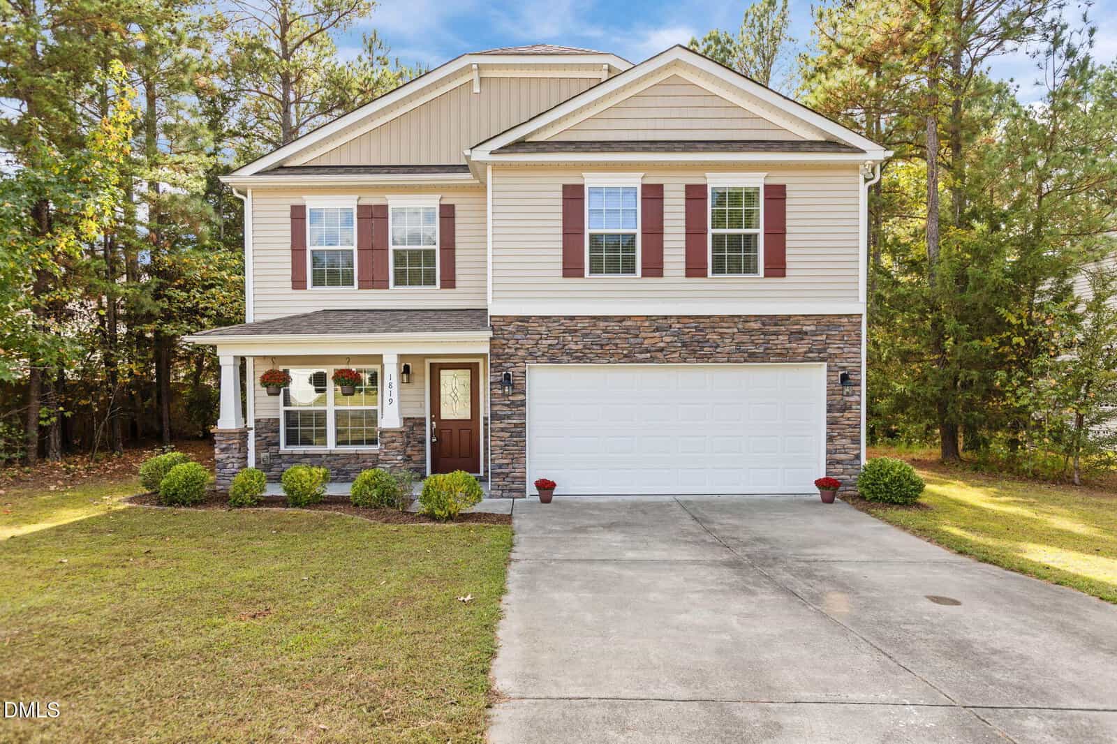 Two-story suburban house at 1819 Fillmore Drive in Creedmoor, featuring tan siding, stone accents, white trim, and a double garage door. The front yard offers trimmed bushes, a green lawn, and concrete driveway with trees in the background.