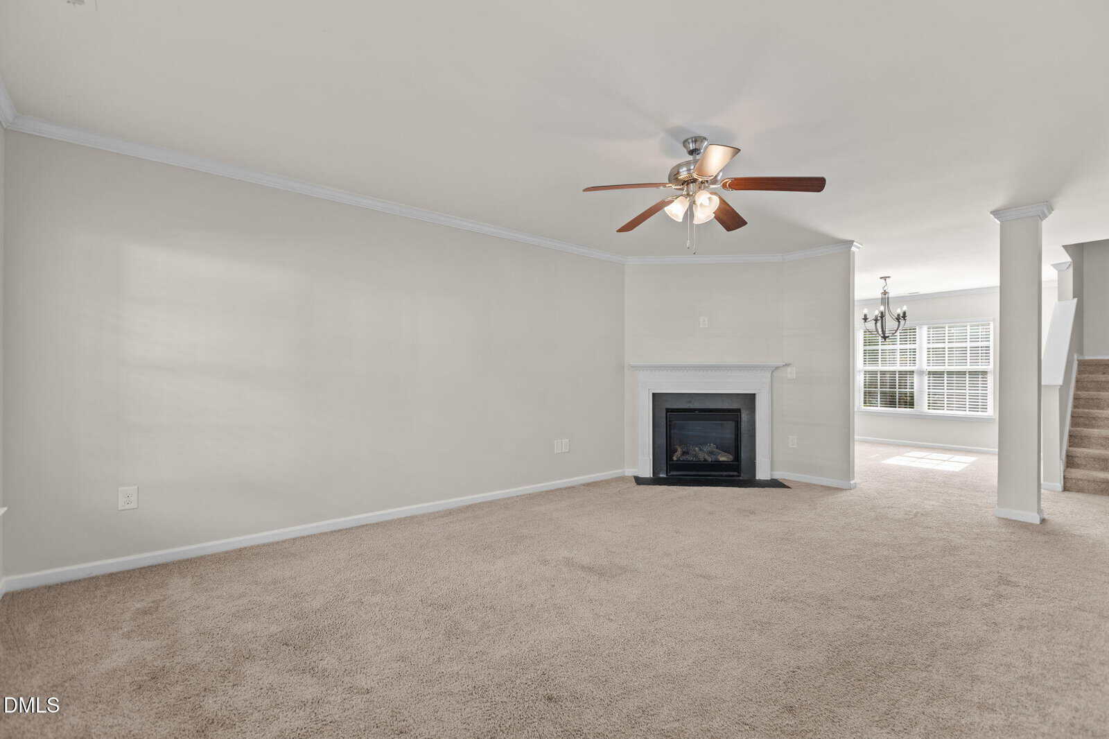 A spacious living room with beige carpet, a ceiling fan with lights, a white fireplace in the corner, light gray walls, and an open view into a dining area with stairs leading up on the right side.