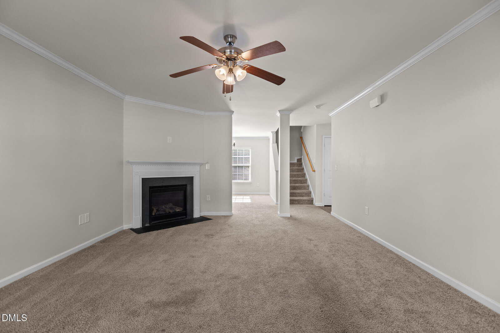 A carpeted living room with beige walls, a ceiling fan with lights, a white fireplace, a window at the back, and a staircase leading upstairs. The room is unfurnished and has white trim.