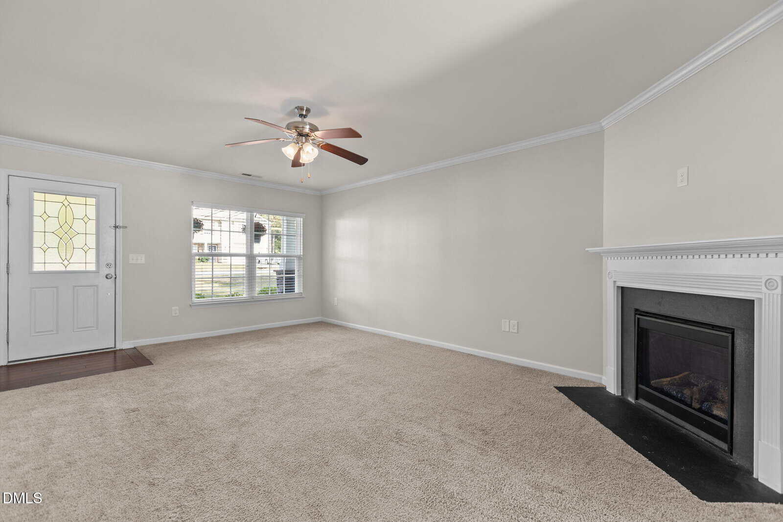 A living room with beige carpet, light-colored walls, a ceiling fan with lights, a window with blinds, a white front door with decorative glass, and a white fireplace with a black hearth.