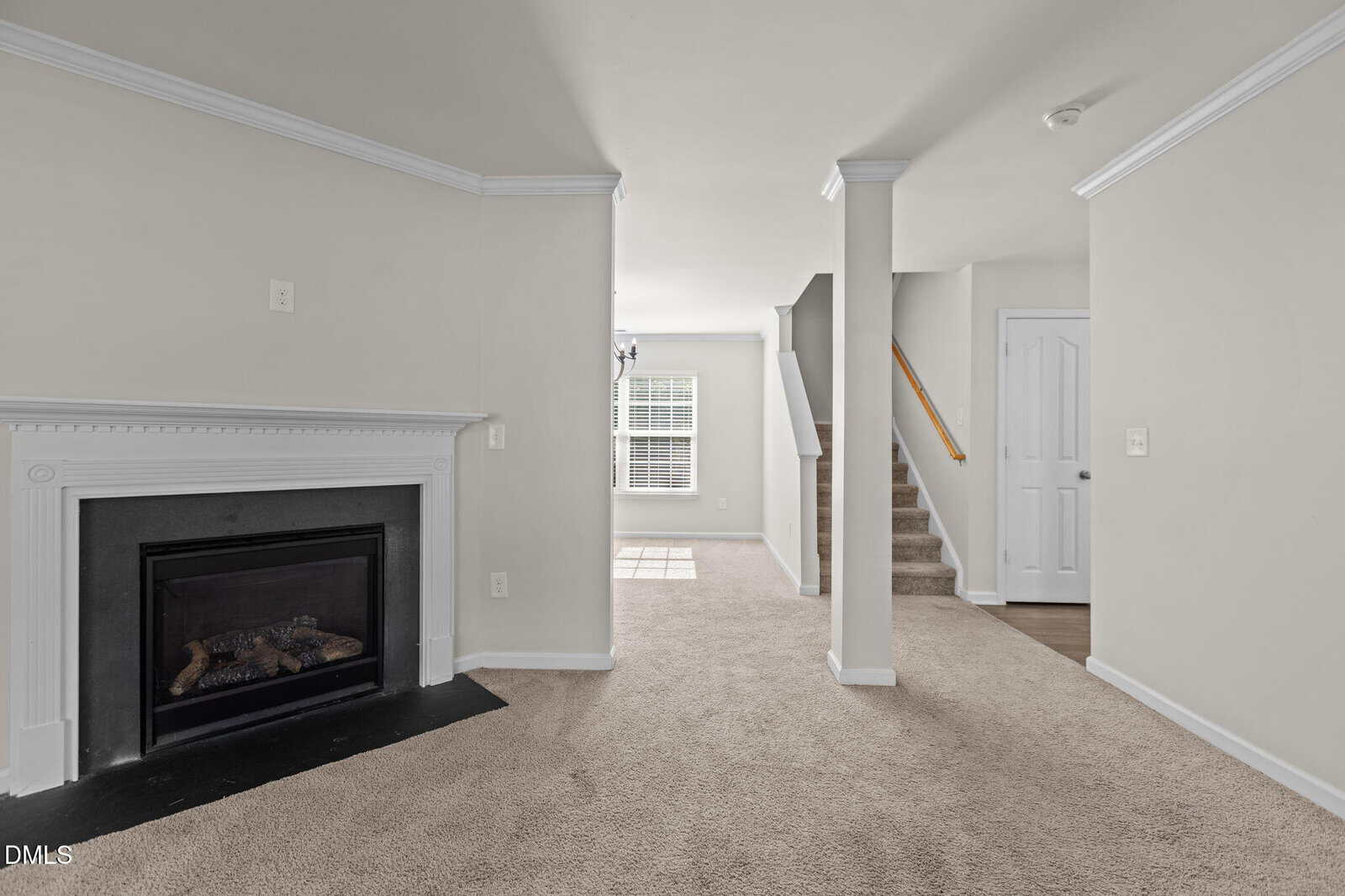 A room with beige carpet, a white fireplace on the left, a staircase with a wooden handrail on the right, and a window in the background letting in natural light. Walls and trim are painted white.