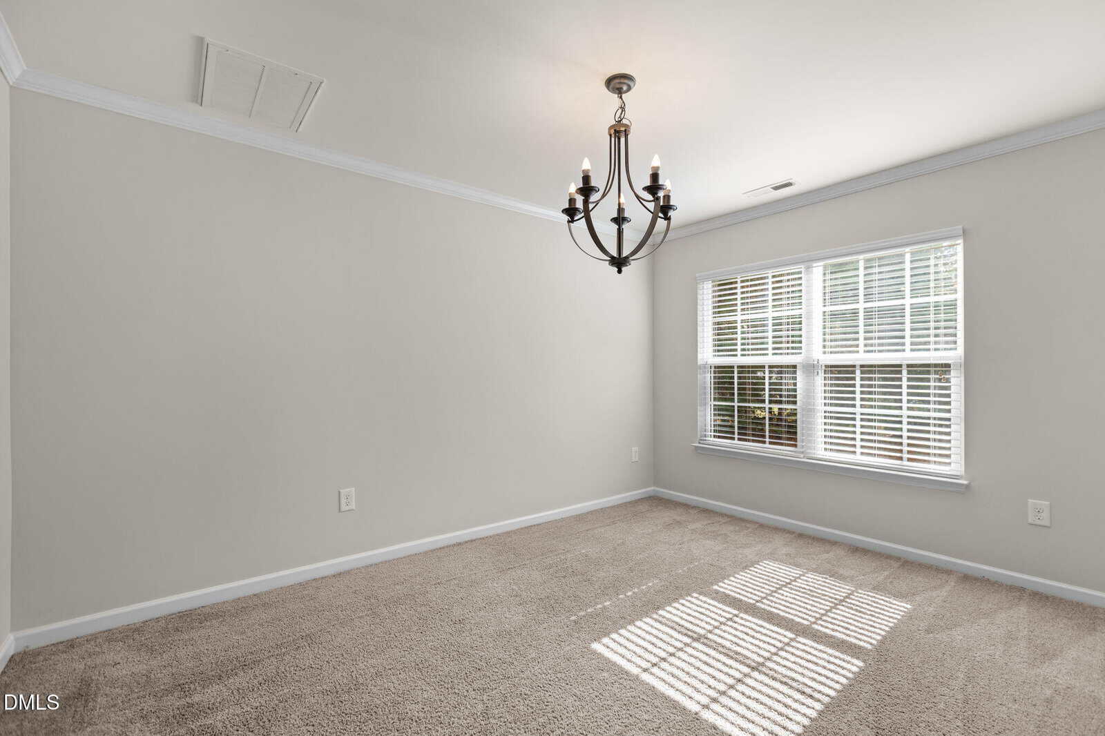 Empty room with beige carpet, light gray walls, white trim, a black metal chandelier on the ceiling, and a large window with white blinds letting in natural light.