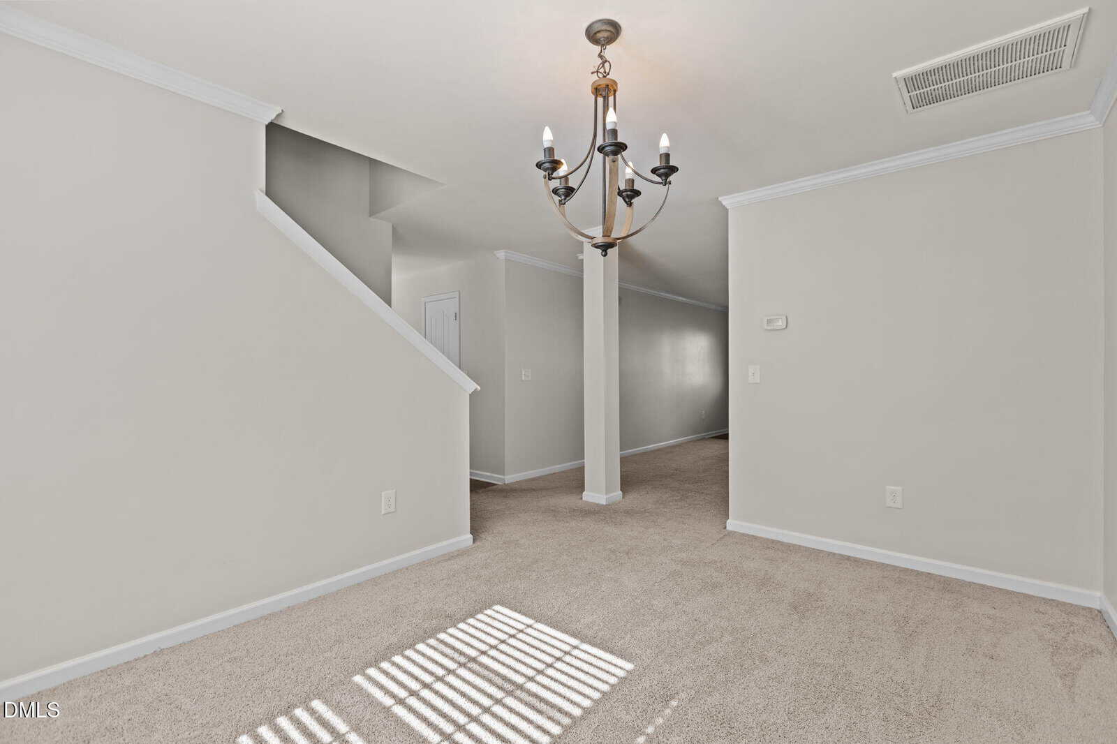 A room with beige carpet, white walls, and a modern black chandelier. An angled staircase with a partial wall leads upward. Sunlight casts window shadows on the floor. Visible door and air vent on the ceiling.