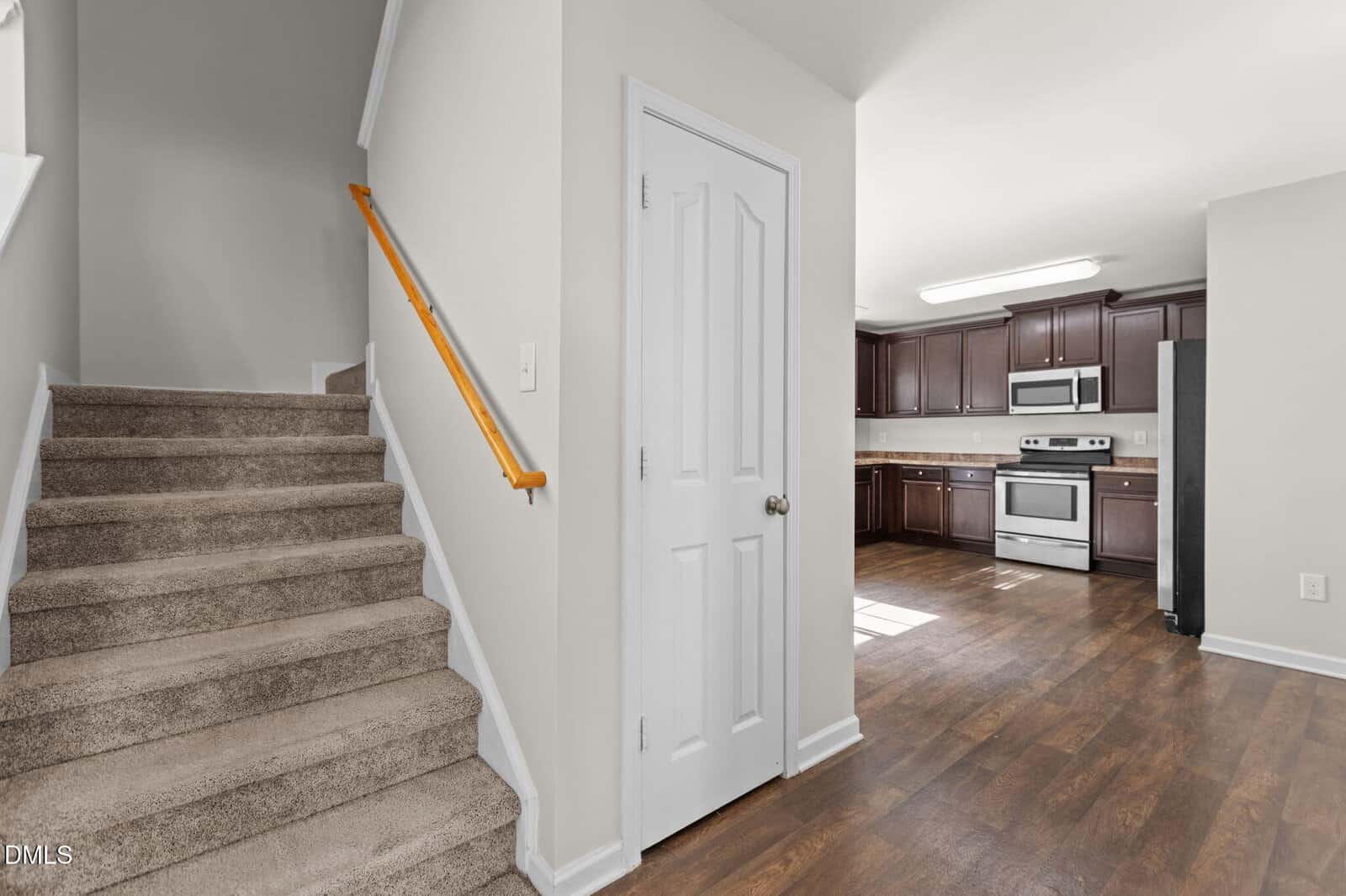 A carpeted staircase with a wooden handrail is on the left, adjacent to a white closet door. An open kitchen with dark cabinets, stainless steel appliances, and wood flooring is visible on the right.