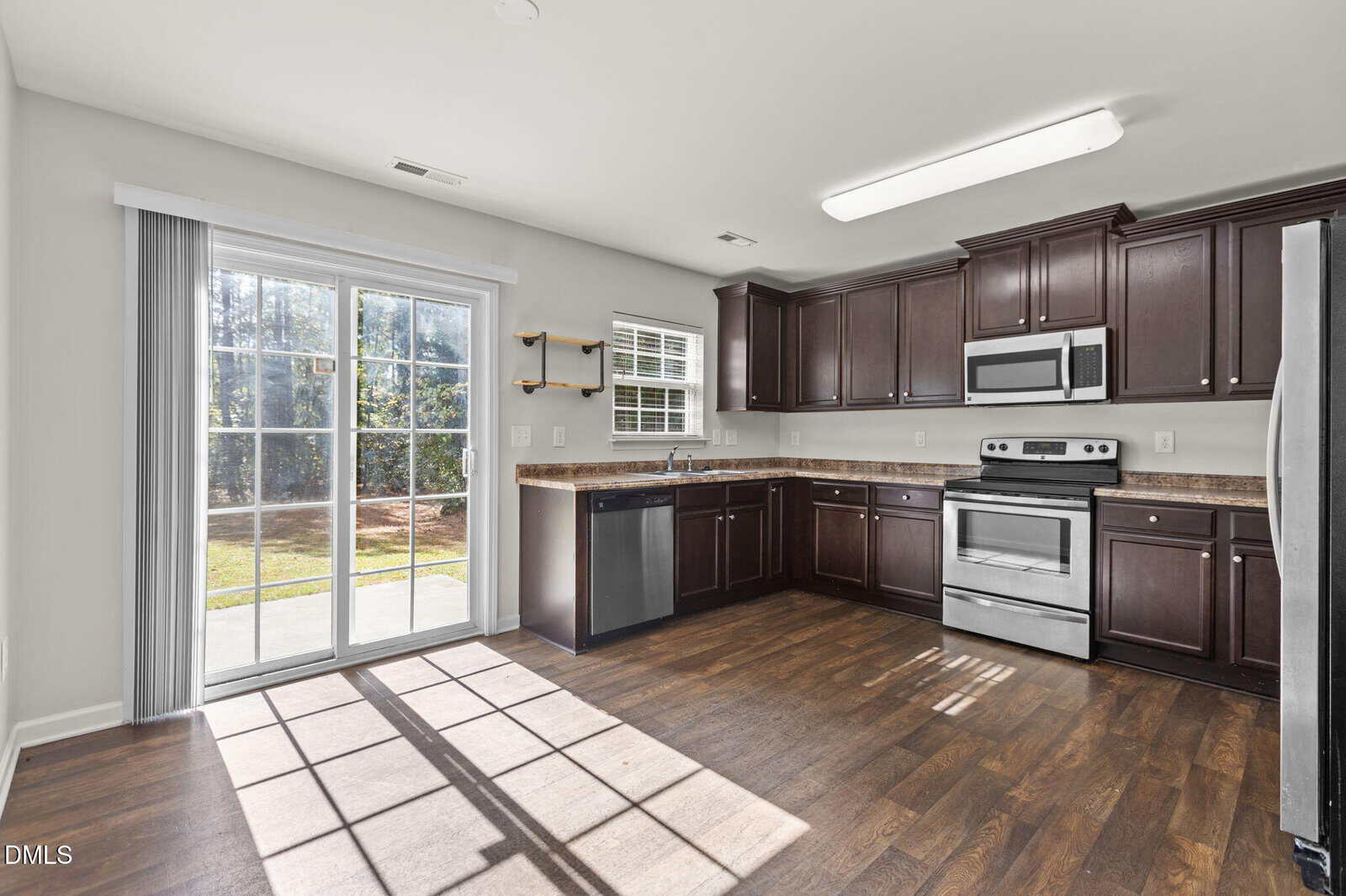 A modern kitchen with dark wood cabinets, stainless steel appliances, brown countertops, and wood flooring. Large sliding glass doors let in natural light, and there is a window above the sink.