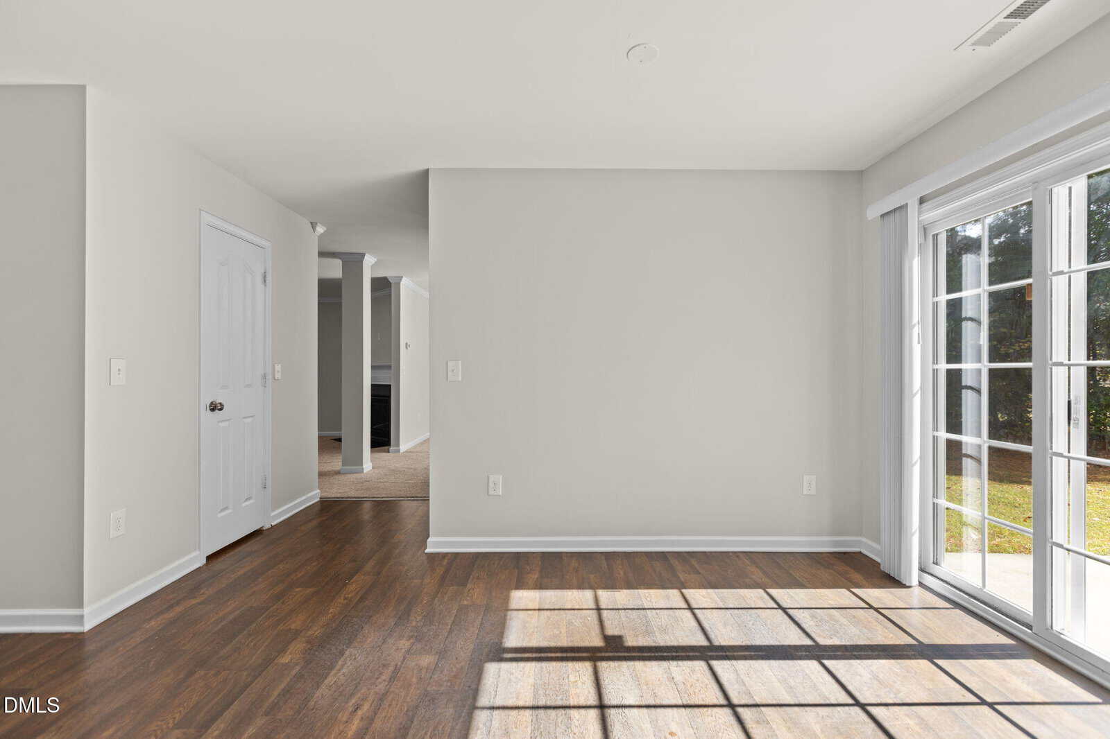 A bright, empty room with wood floors, white walls, a closed white door, and large sliding glass doors letting in sunlight and showing a view of a green yard outside.