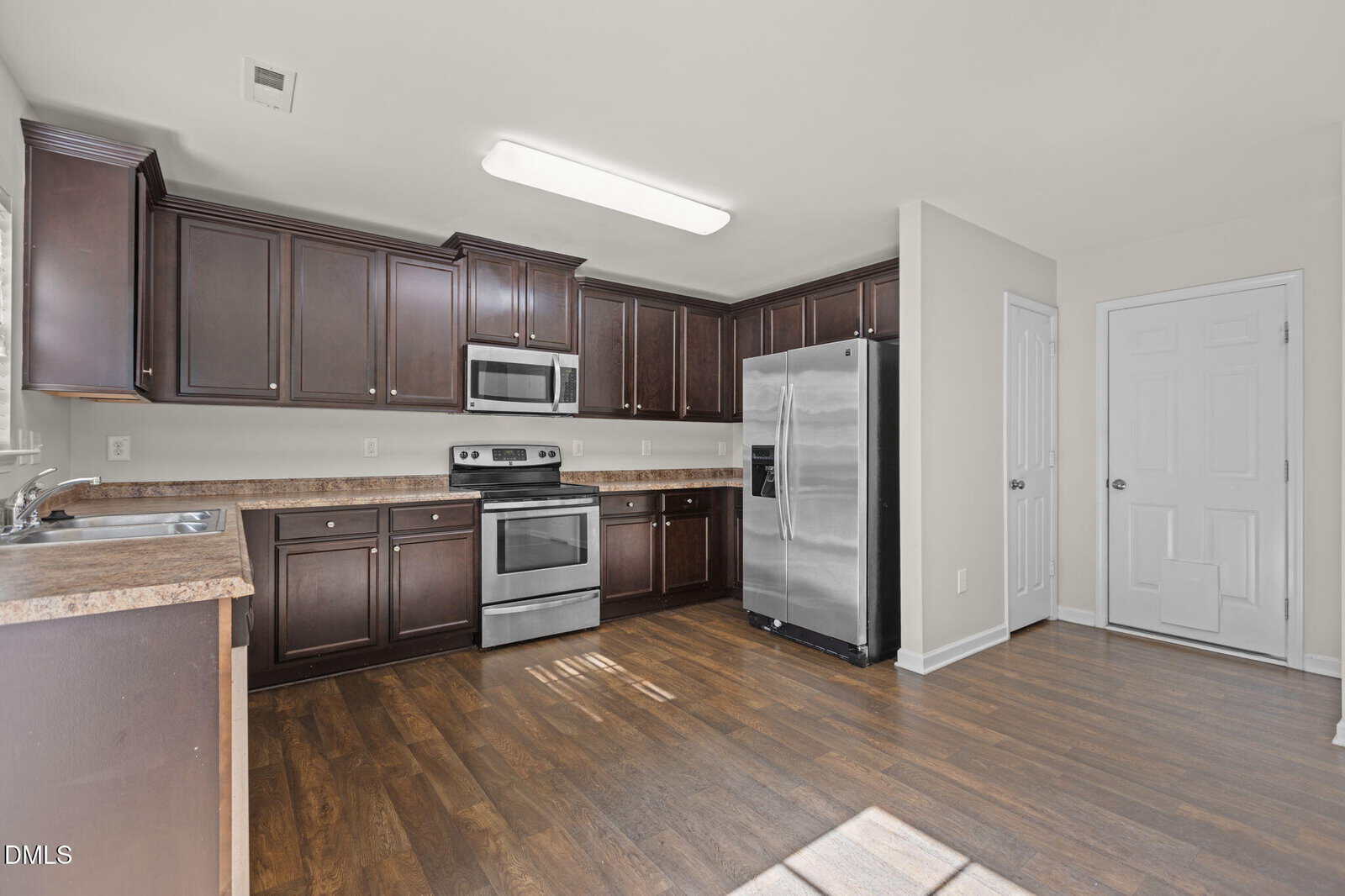 Modern kitchen with dark wood cabinets, stainless steel appliances, light countertops, and wood flooring. A large refrigerator, oven, and microwave are against one wall. White doors and walls complete the clean look.